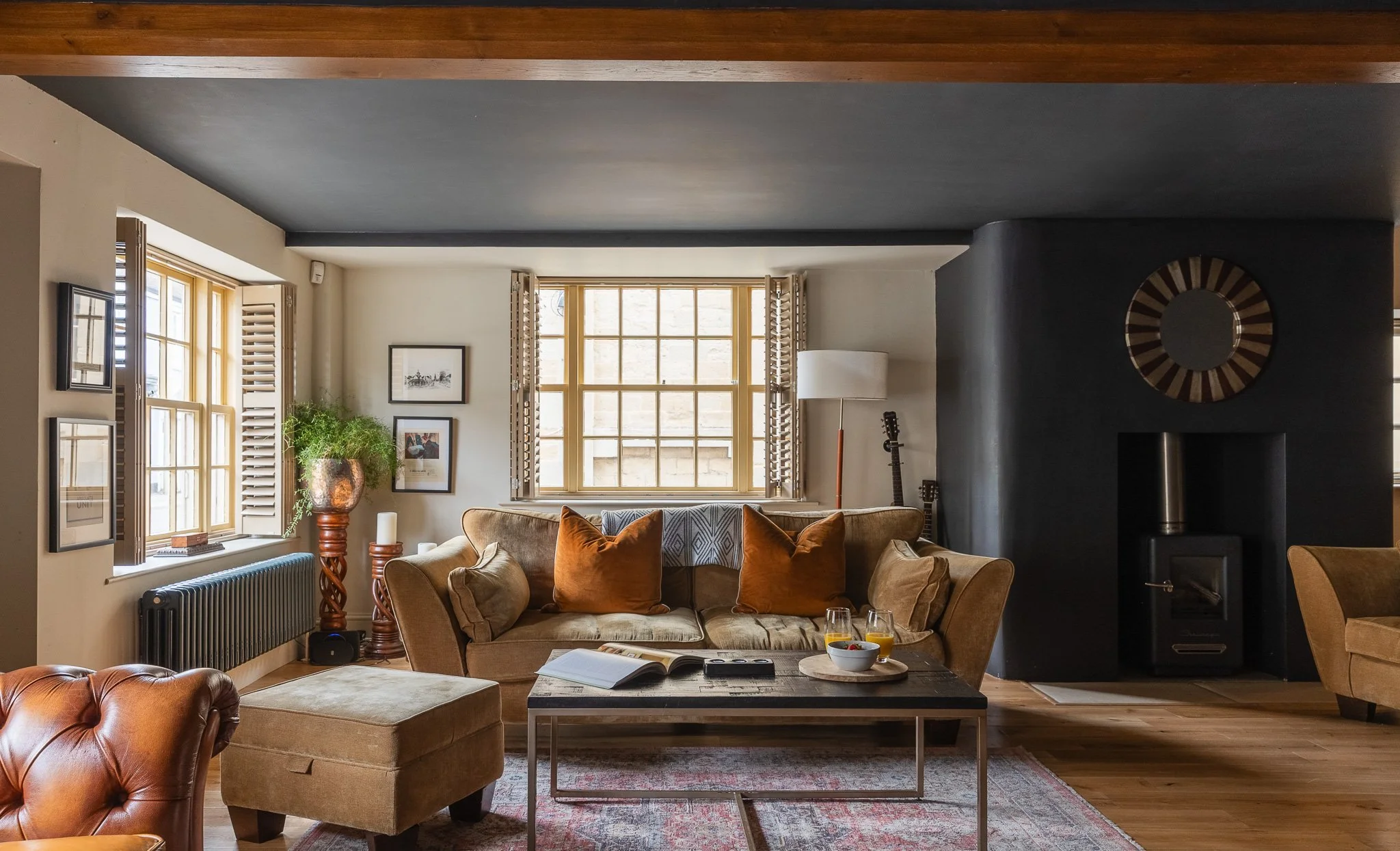 Living room with beige sofa, brown and beige pillows, coffee table with books and drinks, window with wooden shutters, plants, and artwork, black fireplace, armchair, and guitars in the background.