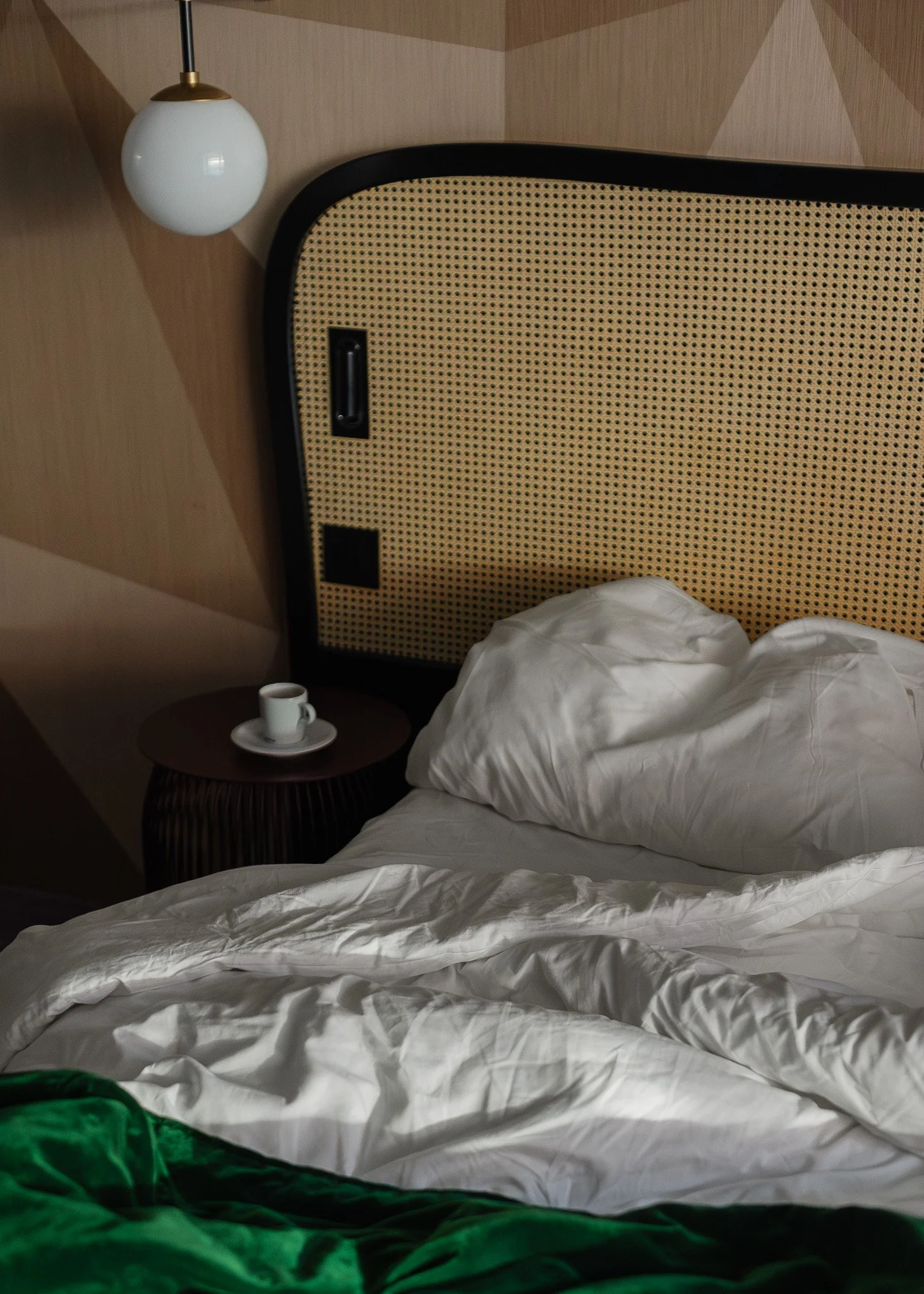 Unmade bed with white sheets and pillows, a nightstand with a cup and saucer, and a wall-mounted lamp in a modern bedroom.