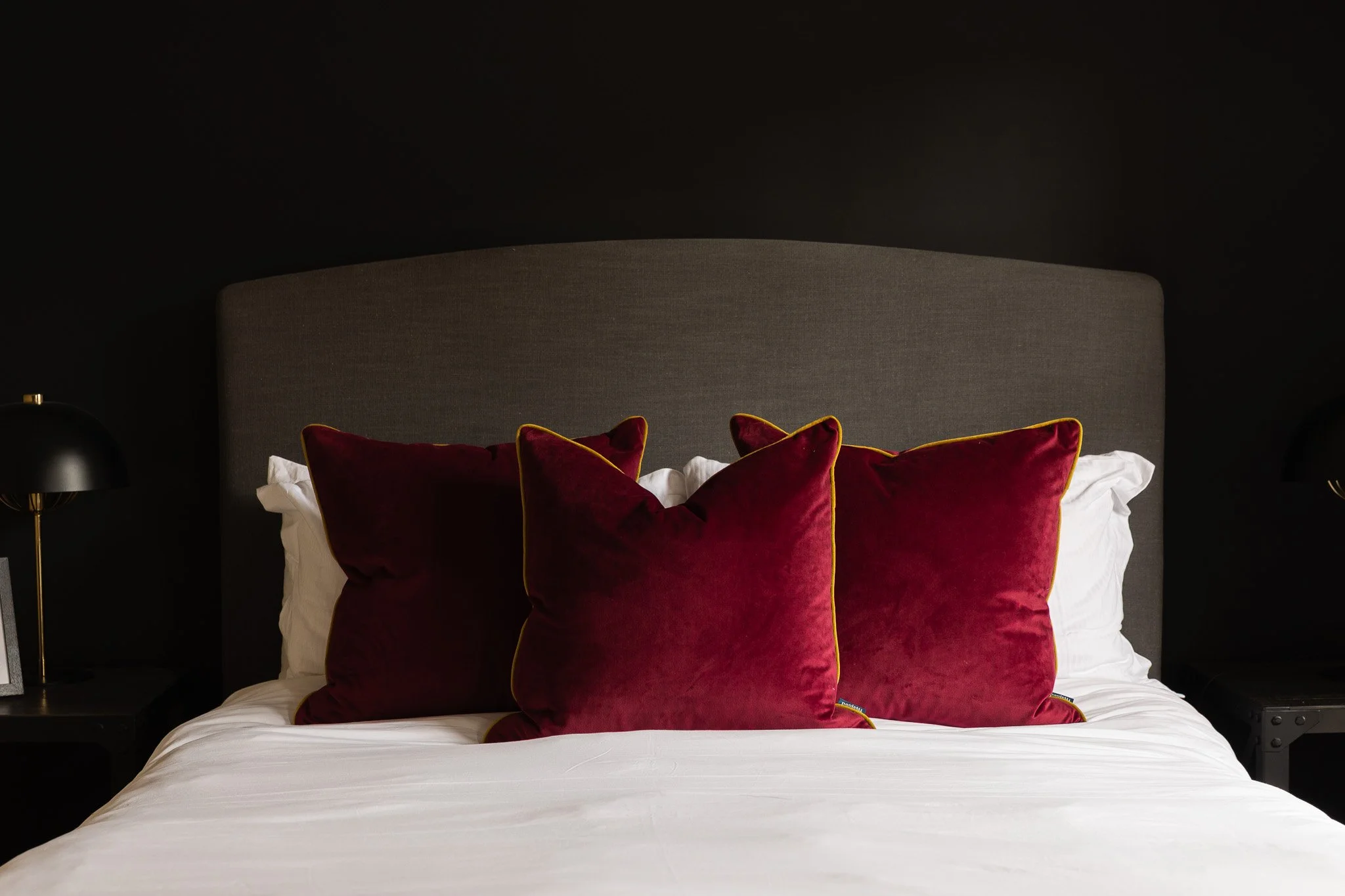 A bed with a dark gray upholstered headboard, white bedsheets, and three dark red velvet pillows with yellow piping, set against a dark wall.