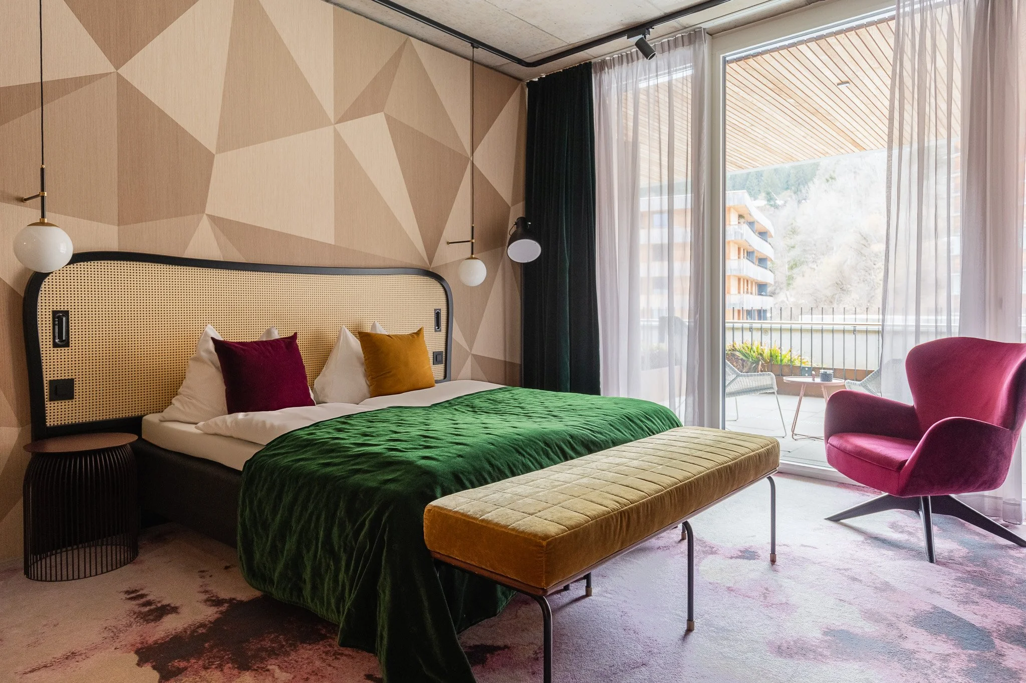 Modern hotel room with a bed that has white, burgundy, and mustard pillows, a green blanket, and a beige upholstered bench. There are hanging pendant lights, a geometric patterned wall, a large window with sheer and dark curtains, a pink armchair, an