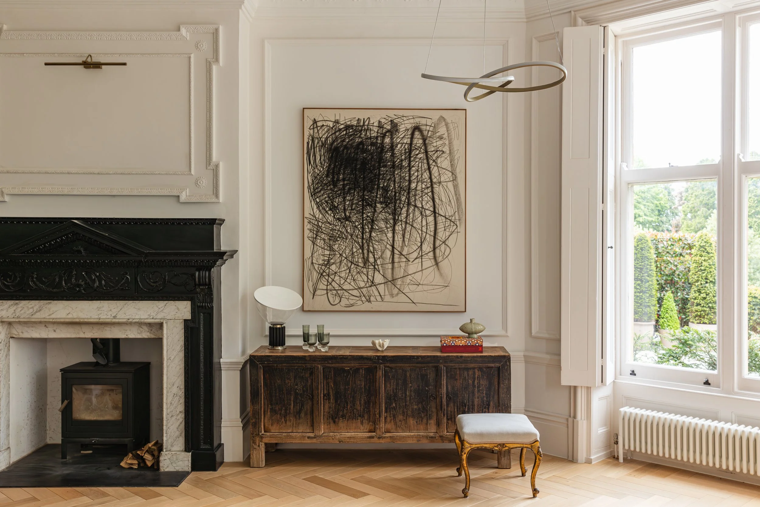 A living room with a black fireplace, a vintage wooden sideboard, abstract art on the wall, a small stool, and large windows letting in natural light, overlooking a green garden.