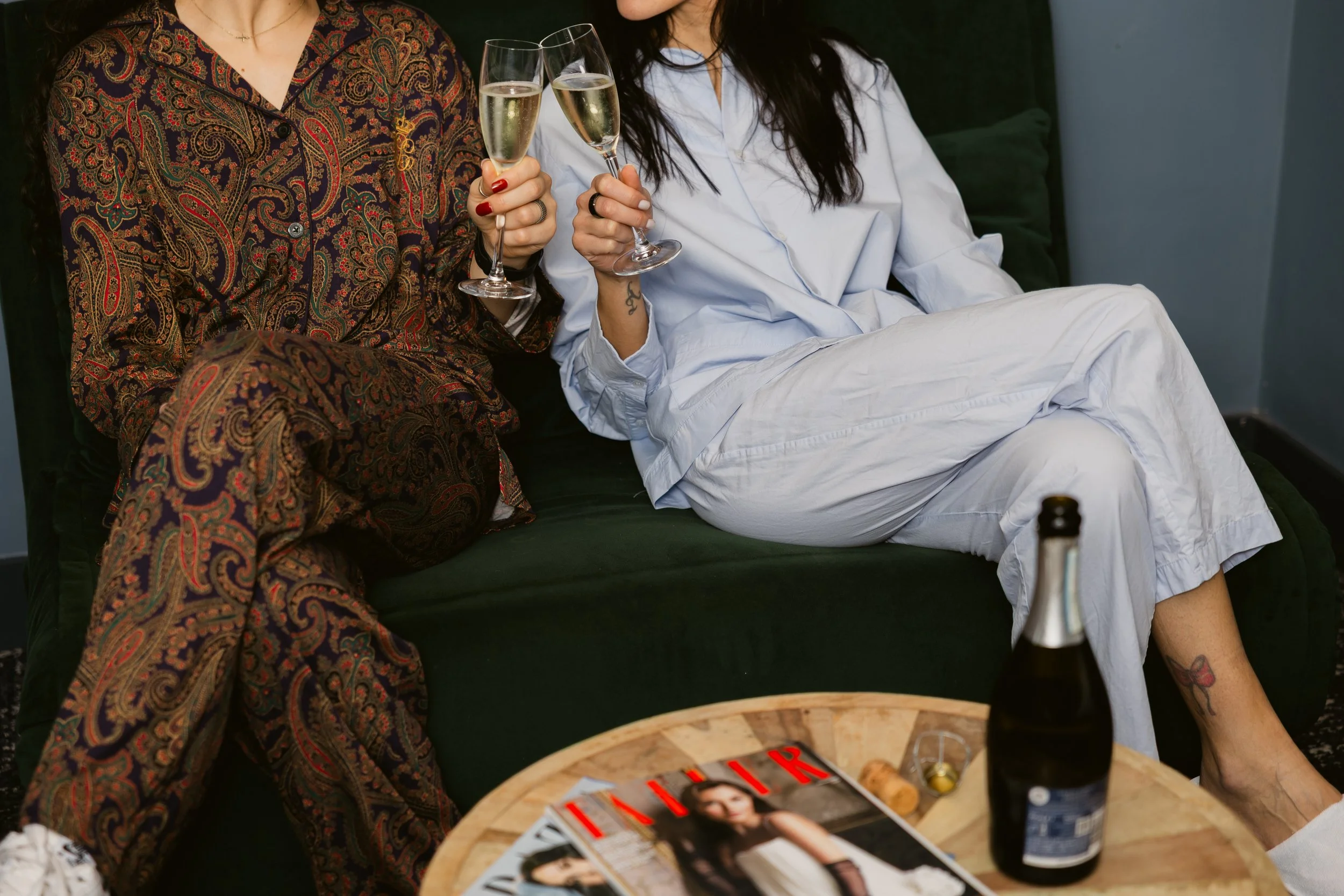 Two women sitting on a dark green couch, holding glasses of champagne, dressed in pajamas or loungewear, with a bottle and magazines on a nearby table.
