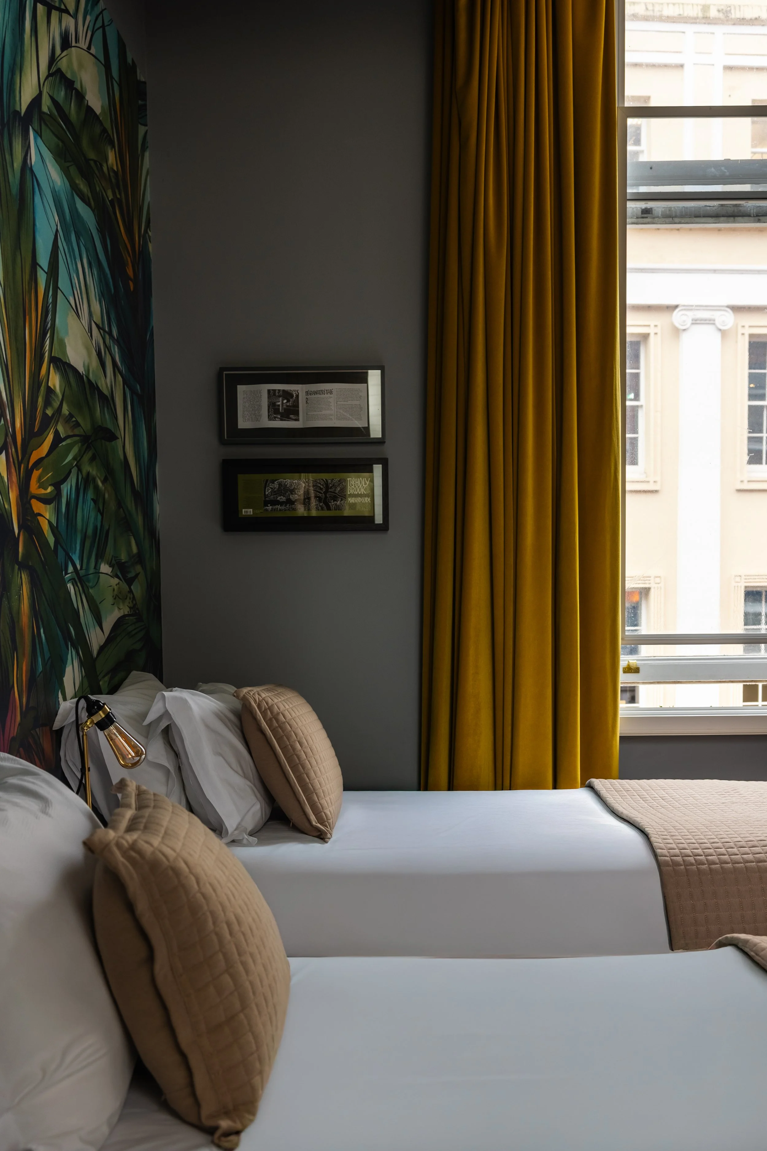 Hotel room with two white beds, beige pillows, and beige blankets, large window with yellow curtains, gray wall with framed pictures, and leafy green mural on the wall.