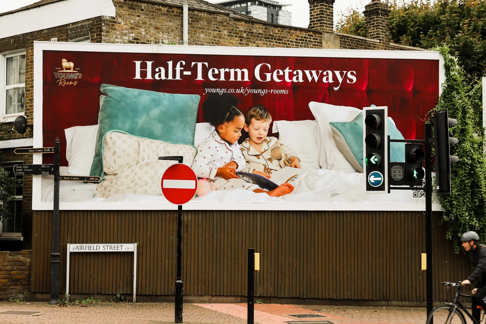 Large billboard advertisement for Young's Rooms featuring a cozy bed with large pillows and two children reading a book together. The banner promotes 'Half-Term Getaways' and includes the website youngs.co.uk/youngs-rooms.