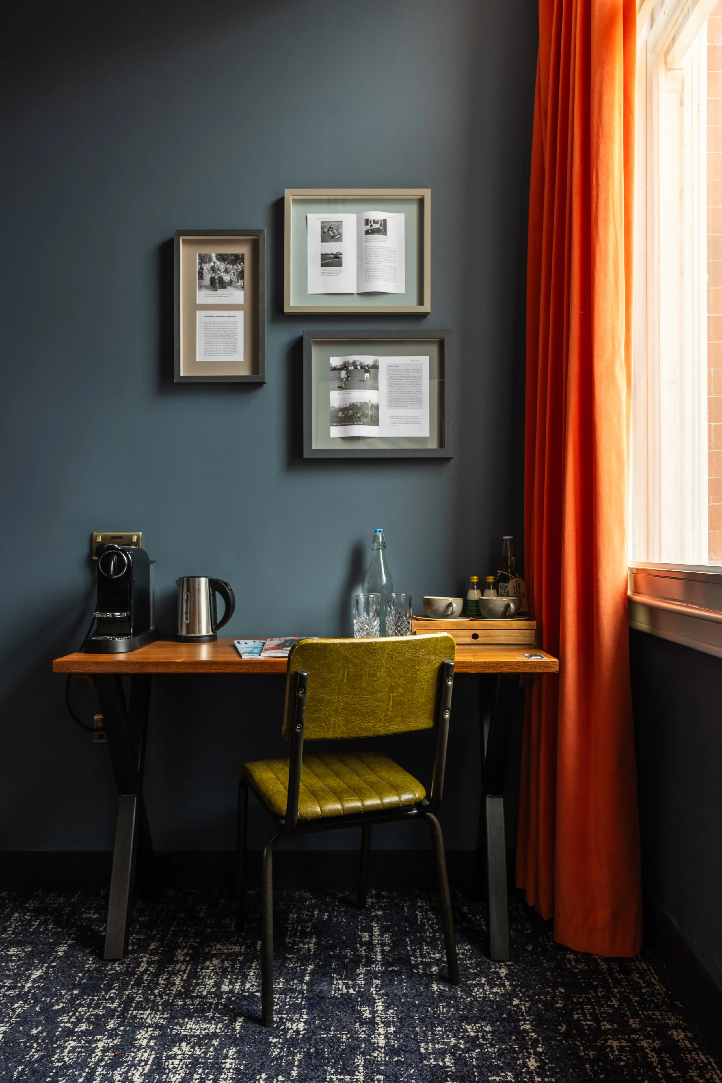 A cozy corner of a room featuring a wooden desk with a coffee machine and electric kettle, an olive green chair, framed photographs and magazines on a dark blue wall, a window with orange curtains, and a patterned rug.