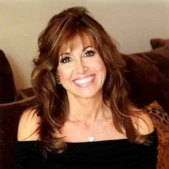 A woman with shoulder-length brown hair smiling, wearing an off-the-shoulder black top, sitting on a brown couch.