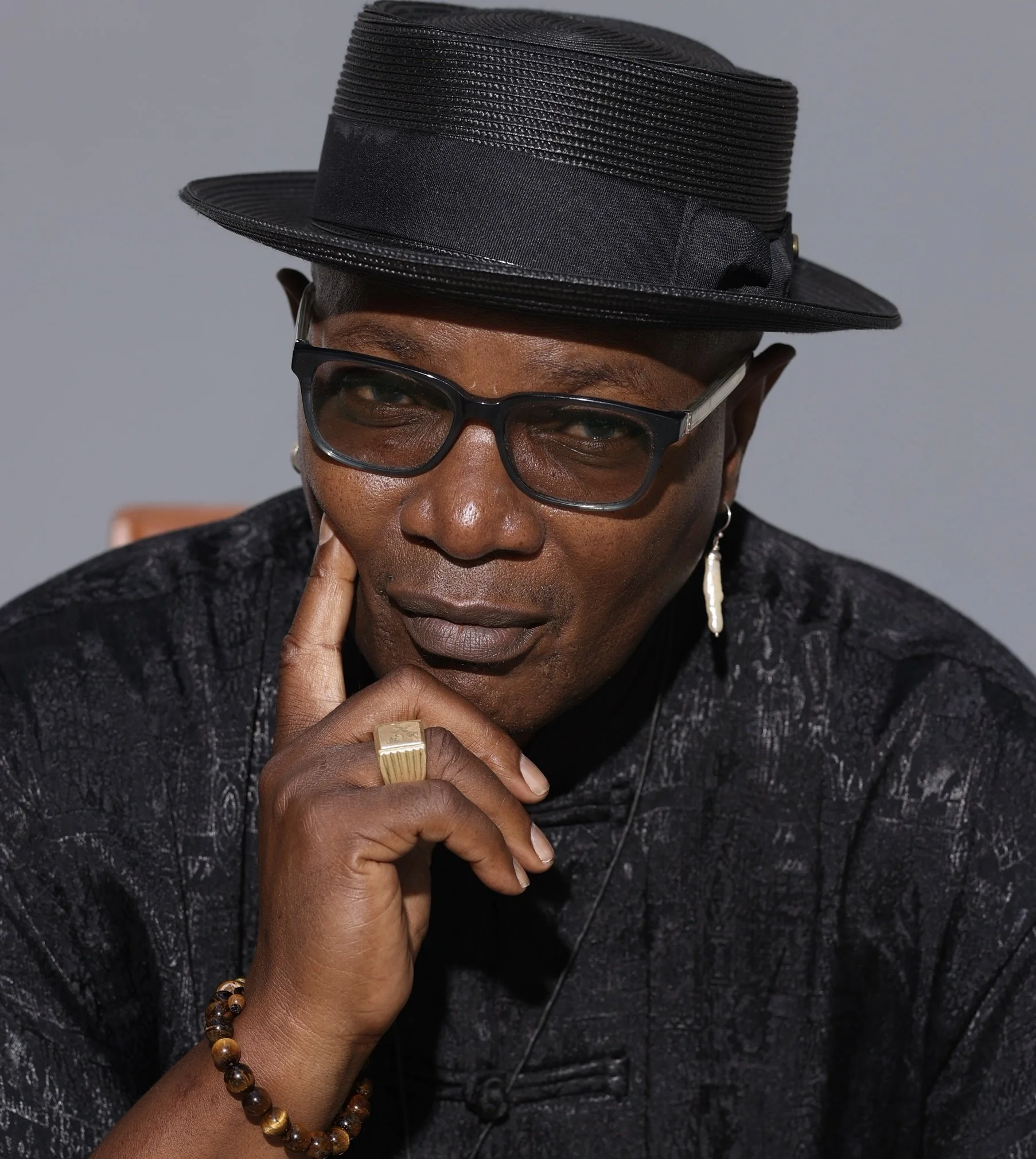 A person wearing a black hat with a wide brim, black glasses, a black textured top, a gold ring, a beaded bracelet, and a pearl earring, striking a thoughtful pose with hand touching face.