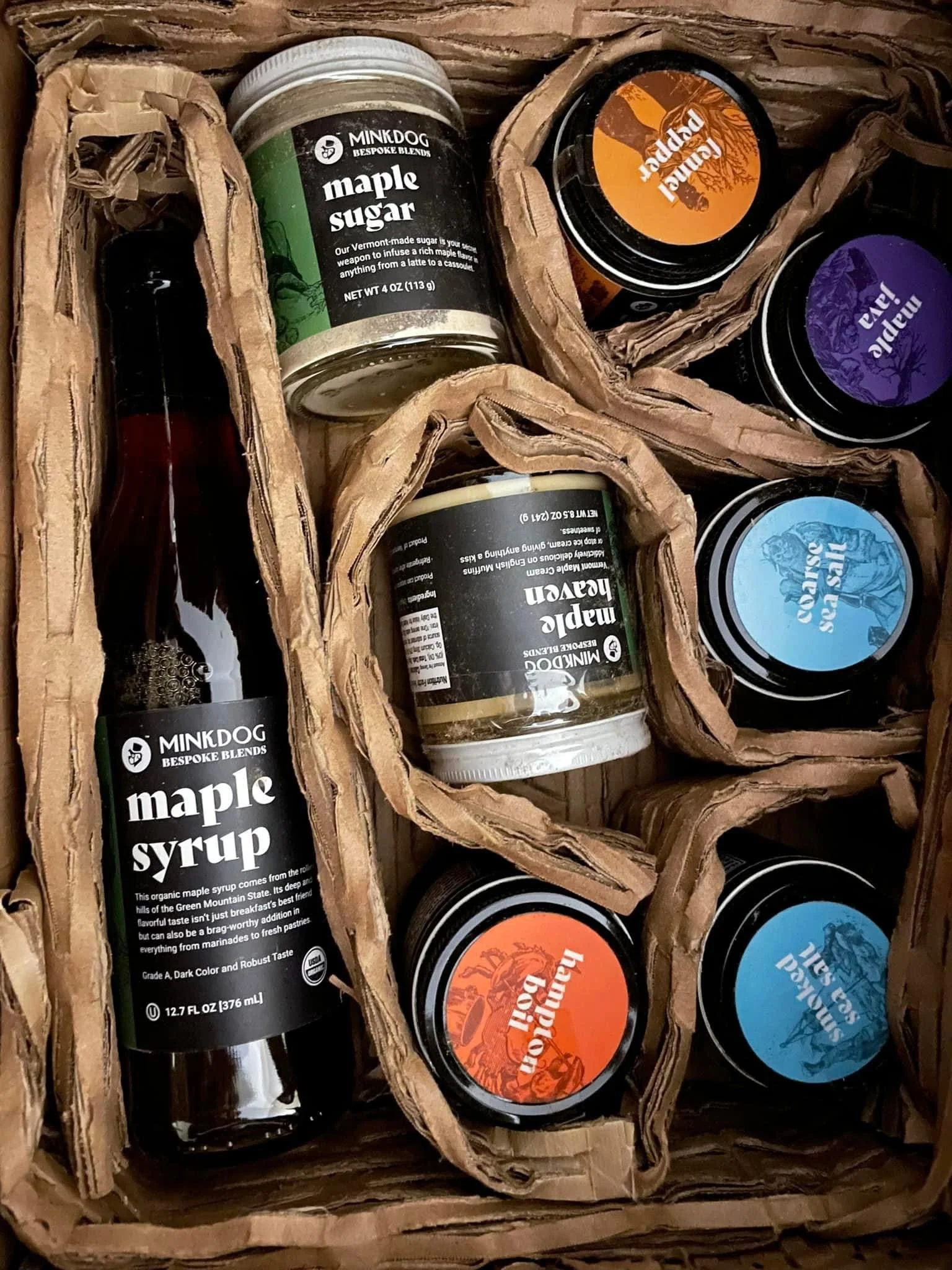 A gift box containing jars and bottles of maple syrup, flavored spreads, and other sweet confections, organized with crumpled brown paper padding.