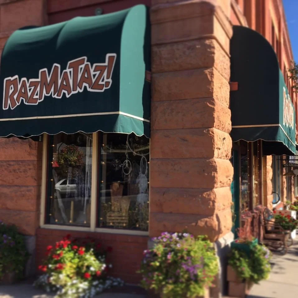 Corner of a brick building with green awnings that have the words "Raz-Ma-Taz!" on them. The windows have plants and a car is visible in the reflection. Flower boxes with colorful flowers are below the windows.