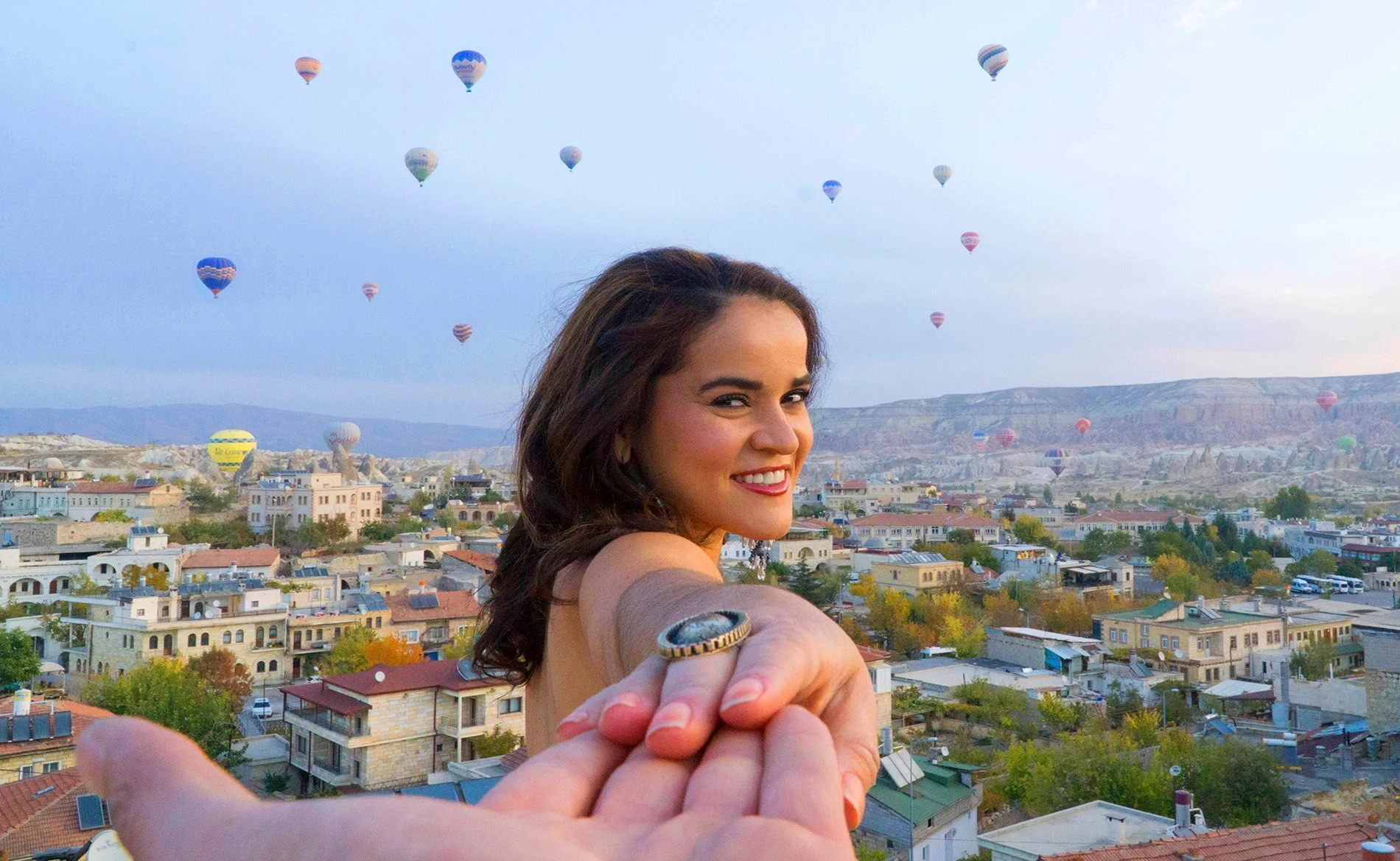 A young woman smiling at the camera, with a cityscape and hot air balloons in the sky behind her.