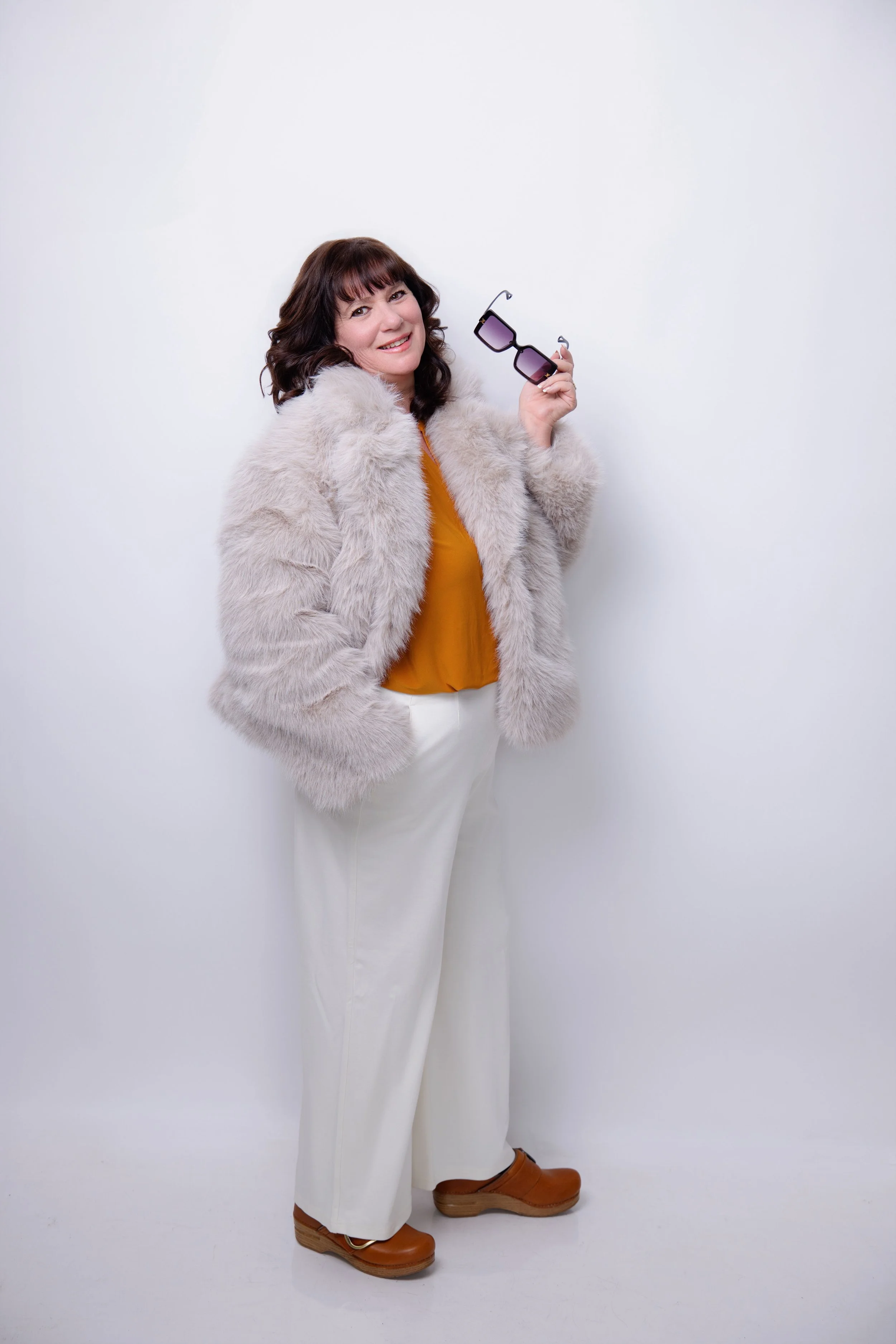 A woman with dark brown hair holding a pair of sunglasses, wearing a light fur coat, a mustard yellow top, white pants, and brown shoes, standing against a plain white background.