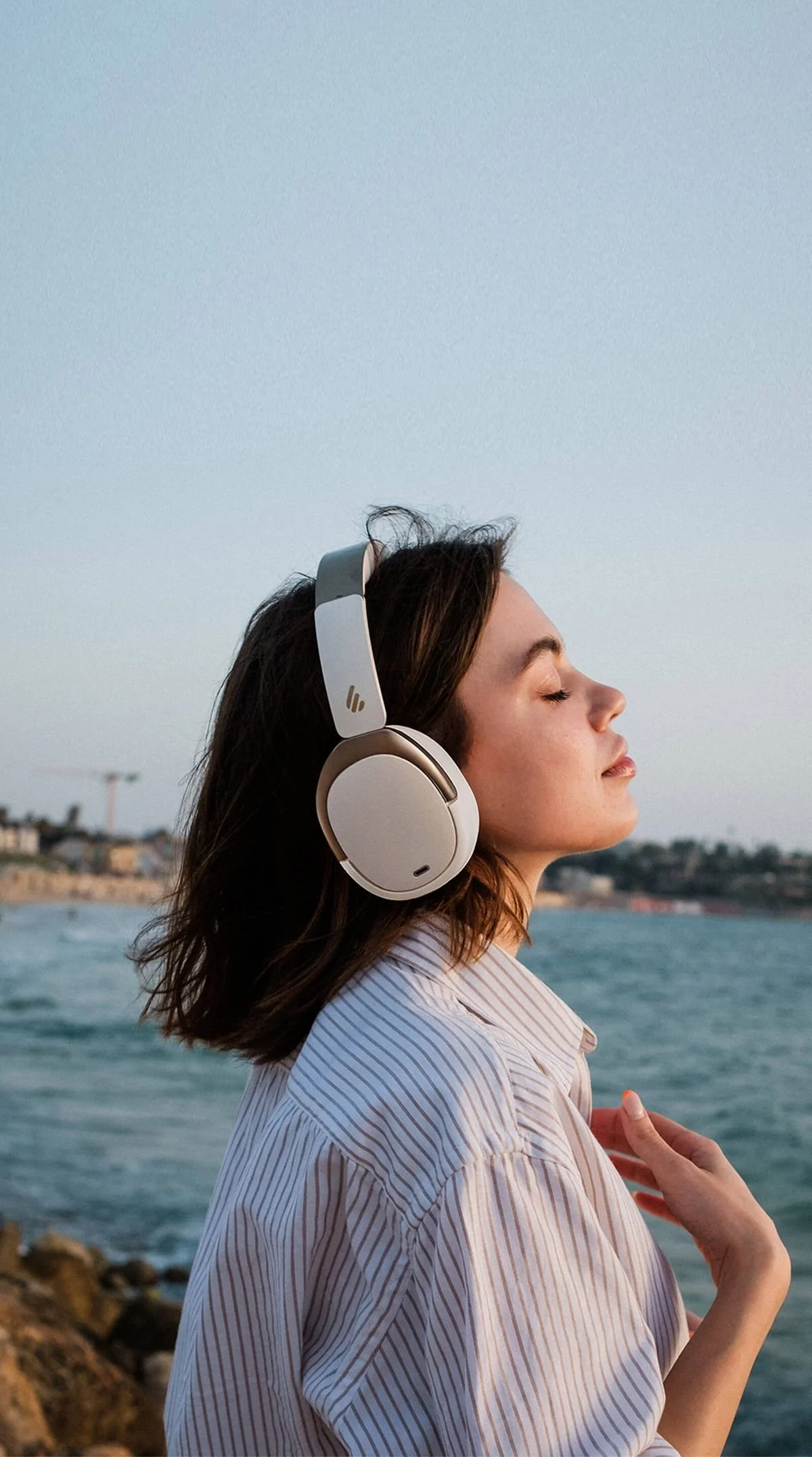 A woman with shoulder-length brown hair wearing large over-ear headphones with her eyes closed, facing a body of water during sunset.