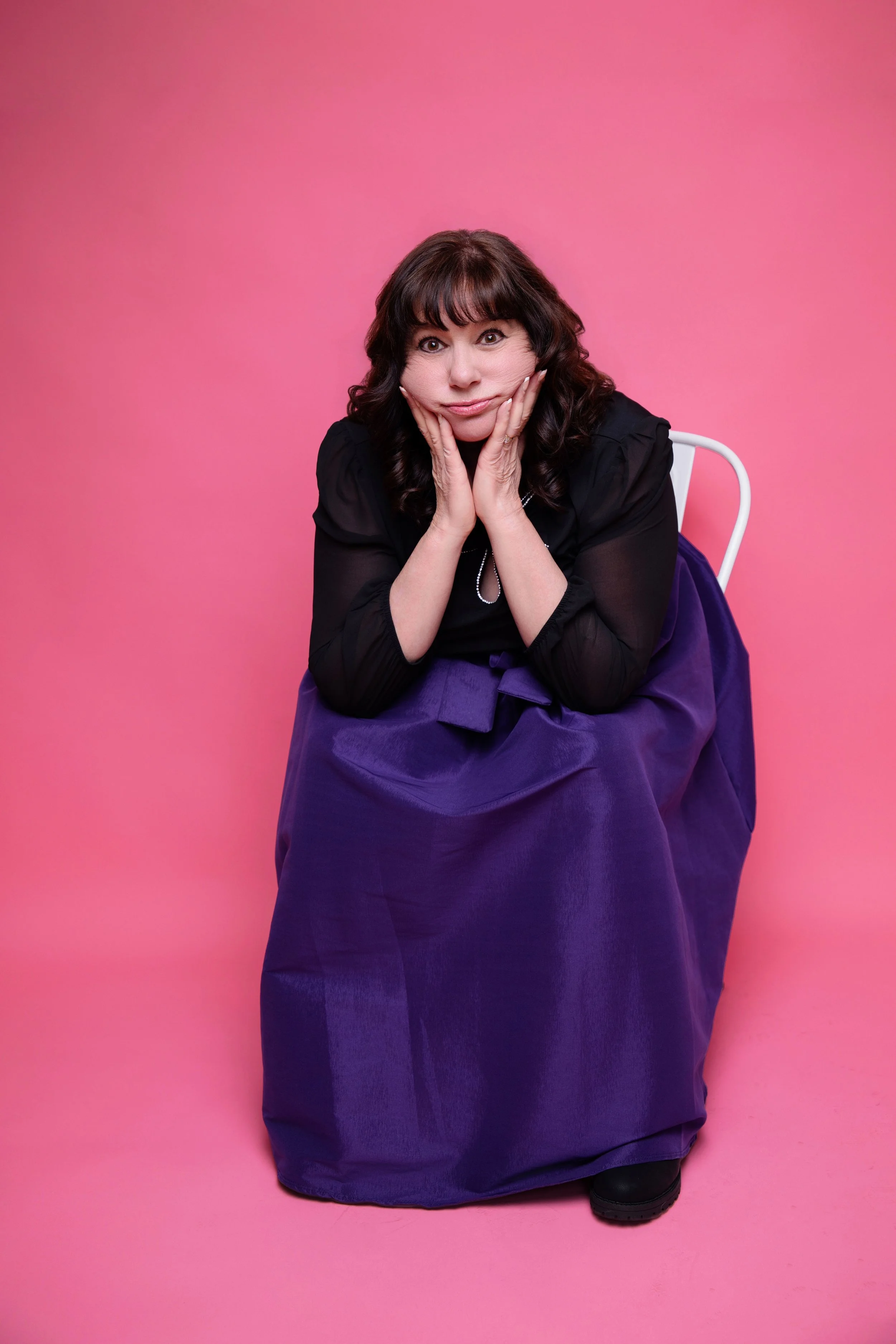 A woman with dark curly hair sitting on a white chair against a pink background. She is wearing a black top and a long purple skirt, resting her chin on her hands in a thoughtful pose.