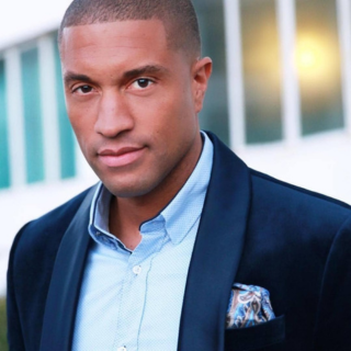 Portrait of a young African American man wearing a navy blazer and light blue shirt, outdoors with a blurred background of windows and lights.