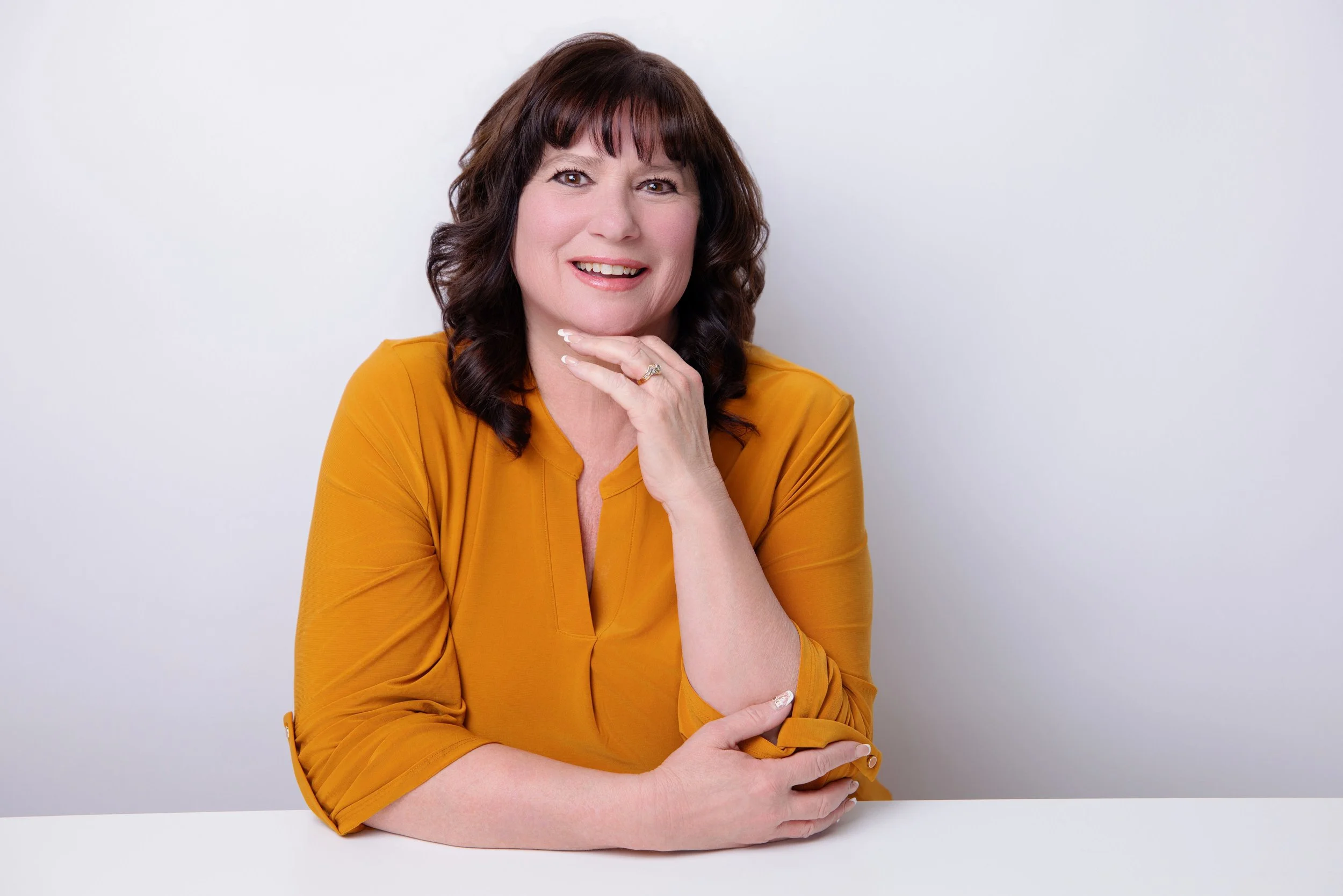 A woman with brown, curly hair wearing a mustard yellow blouse, sitting at a white table, smiling, with her right hand near her face and her left arm resting on the table, against a plain white background.