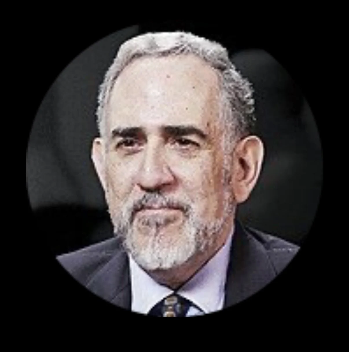 Close-up of a middle-aged man with gray hair and beard, wearing a suit and tie, against a dark background.