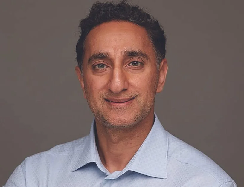A middle-aged man with dark curly hair, light skin, and blue eyes, wearing a light blue collared shirt against a plain gray background.