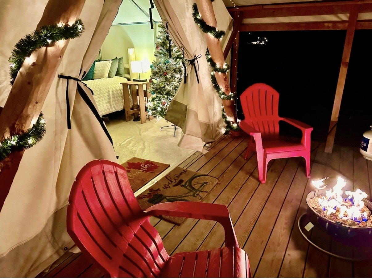 A cozy outdoor porch with wooden flooring, two pink Adirondack chairs, a fire pit with a small fire, and a decorated Christmas tree in the background. The porch is decorated with garland and lights, and there is a glimpse into a bedroom with a bed and a nightstand nearby.