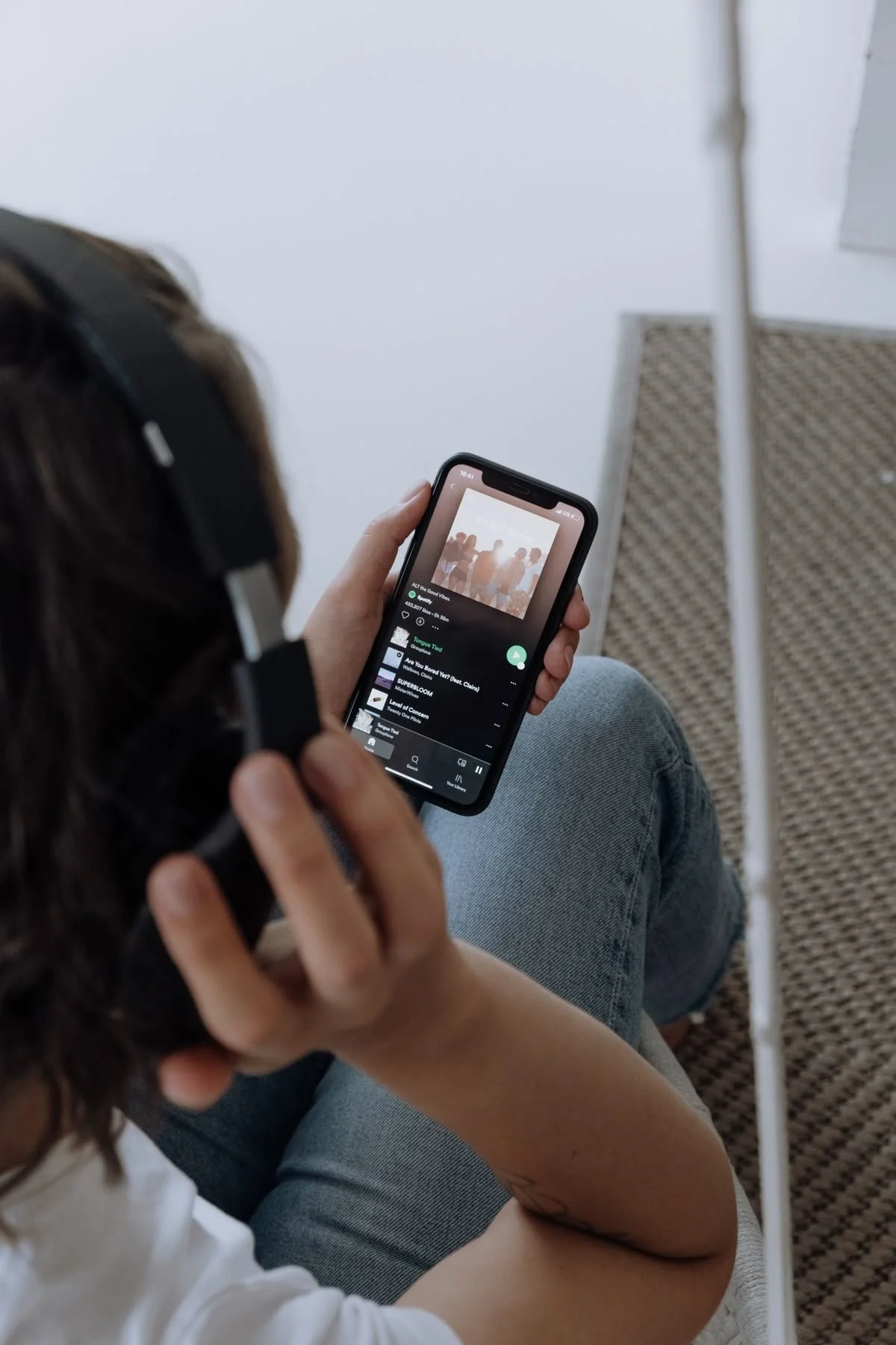 Person sitting with headphones, holding a smartphone, streaming music on Spotify app, with a textured bench and white wall in background.