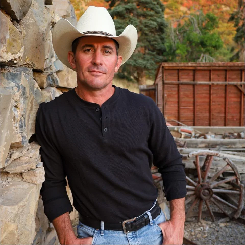 A man wearing a white cowboy hat, black shirt, and jeans leaning against a stone wall outdoors with fall foliage and an old wooden wagon in the background.