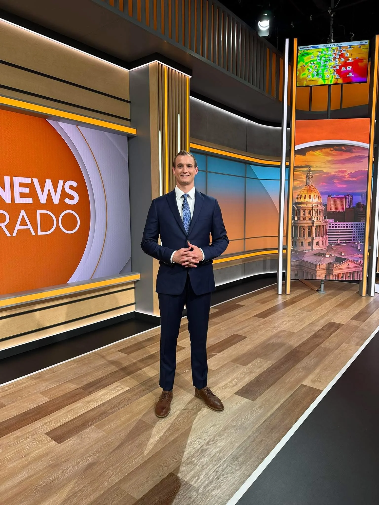 News presenter in a studio with a large screen displaying a cityscape and weather graphics.