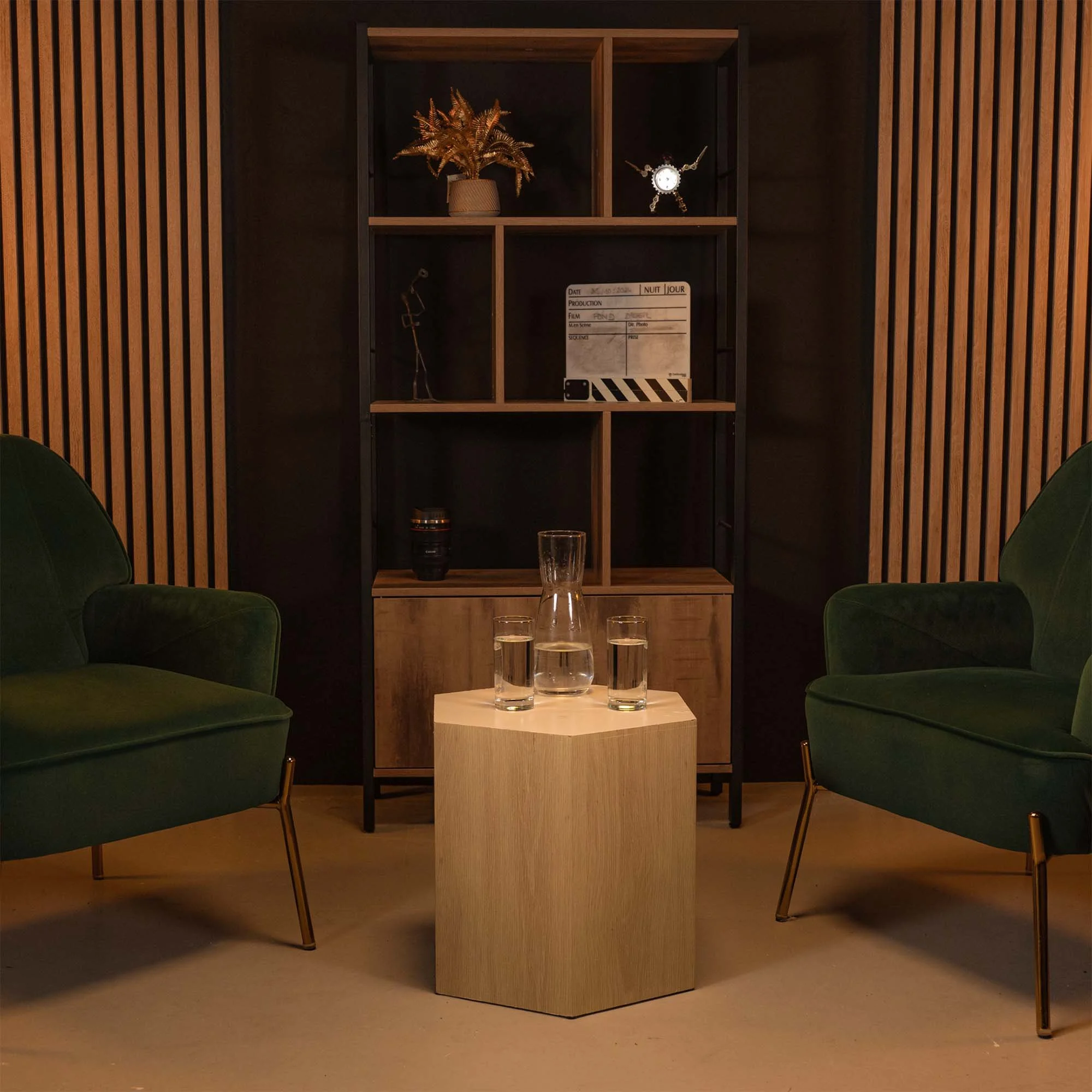 Un salon avec deux fauteuils verts, une table en bois clair avec une carafe et trois verres d'eau, une étagère en bois noire derrière avec des objets décoratifs, un mur en bois avec des lamelles verticales.
