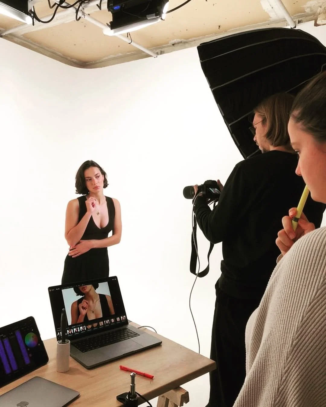 Une séance photo en studio cyclorama avec une femme en costume noir, un photographe avec un appareil photo, et deux assistantes, une tenant un stylo et l'autre une tablette graphique. 