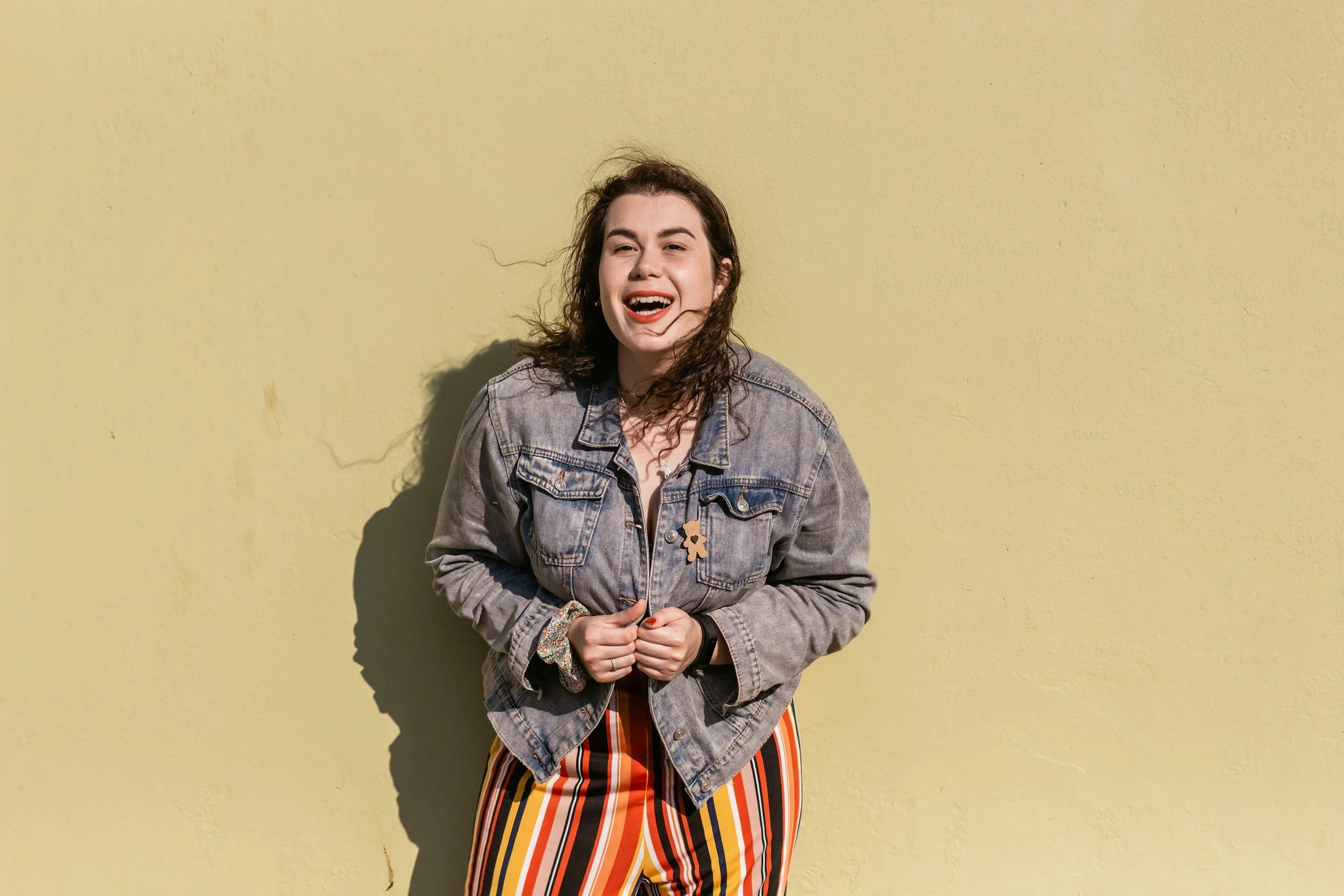 A young female therapist with dark wavy hair wearing a denim jacket and patterned pants standing against a yellow wall, smiling and looking at the camera.