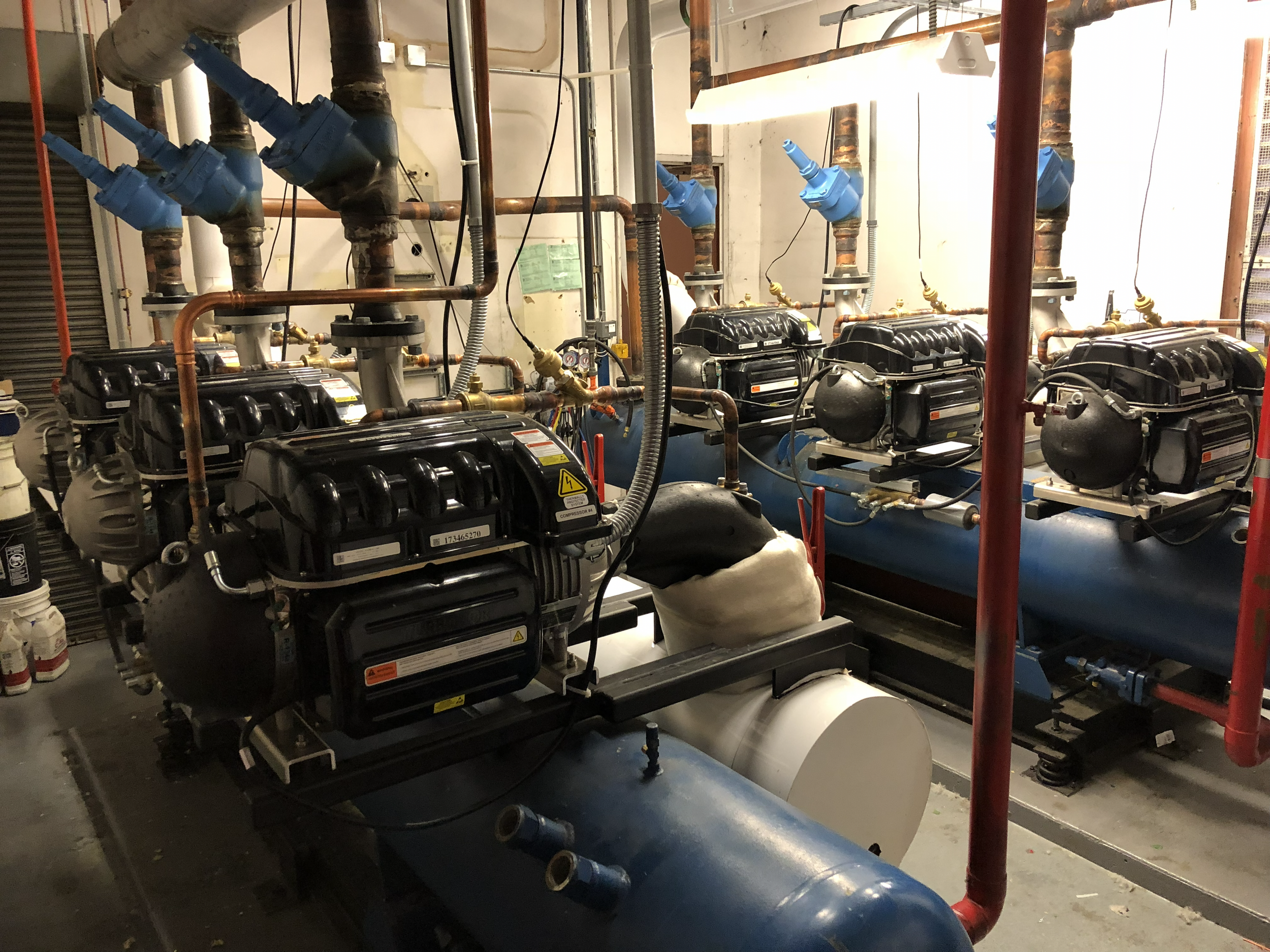 Industrial mechanical room with large blue pipes, black compressors, and copper piping, illuminated by fluorescent lights.