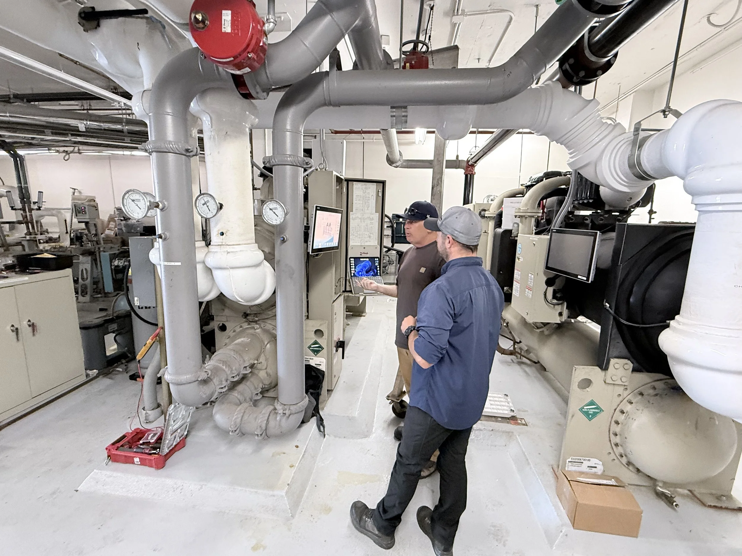 Two workers in blue and gray shirts standing amid industrial pipes and equipment in a mechanical room, with one working on a control panel and the other observing.