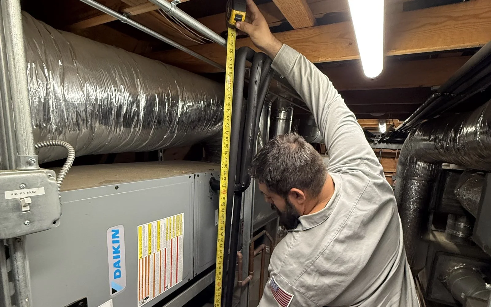 A technician working on an HVAC system in an attic, measuring with a tape measure. Surrounded by ductwork and HVAC equipment.