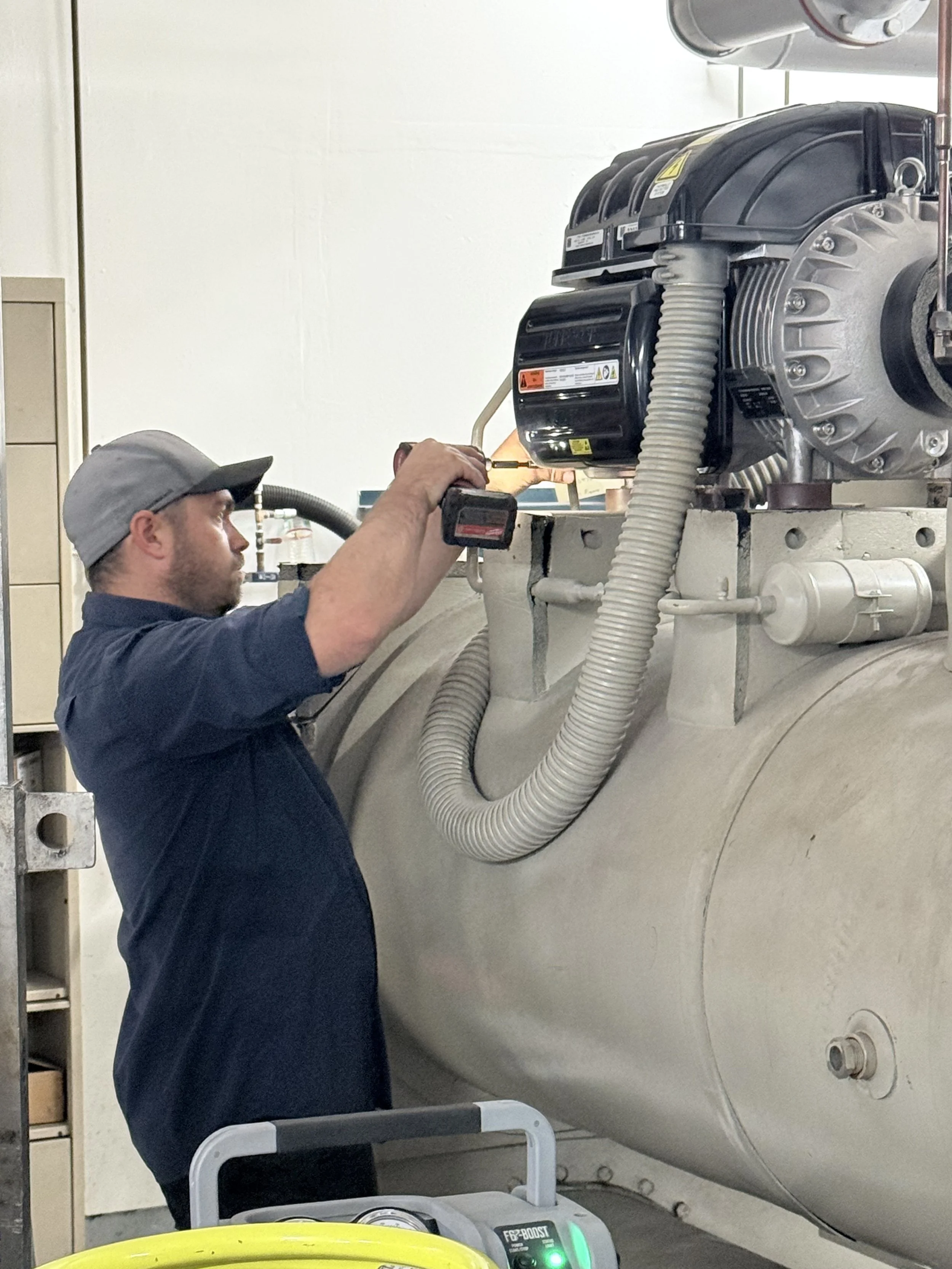 A man wearing a gray cap and glasses is working on a large mechanical or industrial component, possibly a tank or pressure vessel, using a screwdriver or similar tool.