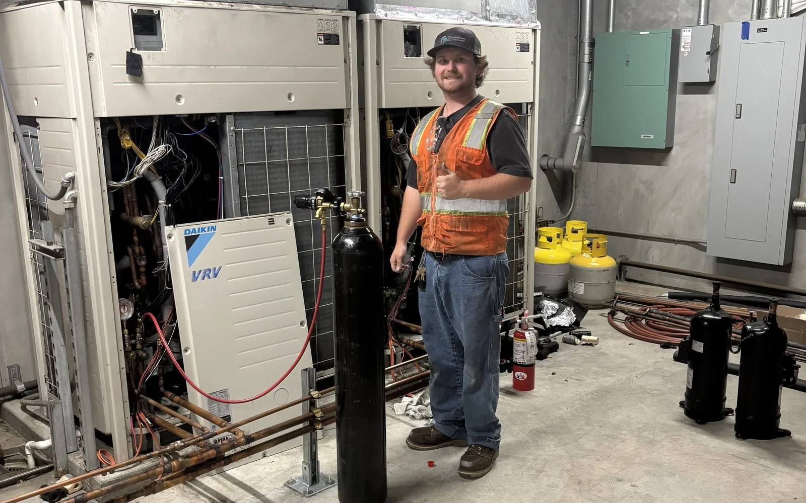 A technician in safety gear working on HVAC equipment with various tools and refrigerant cylinders in an industrial setting.