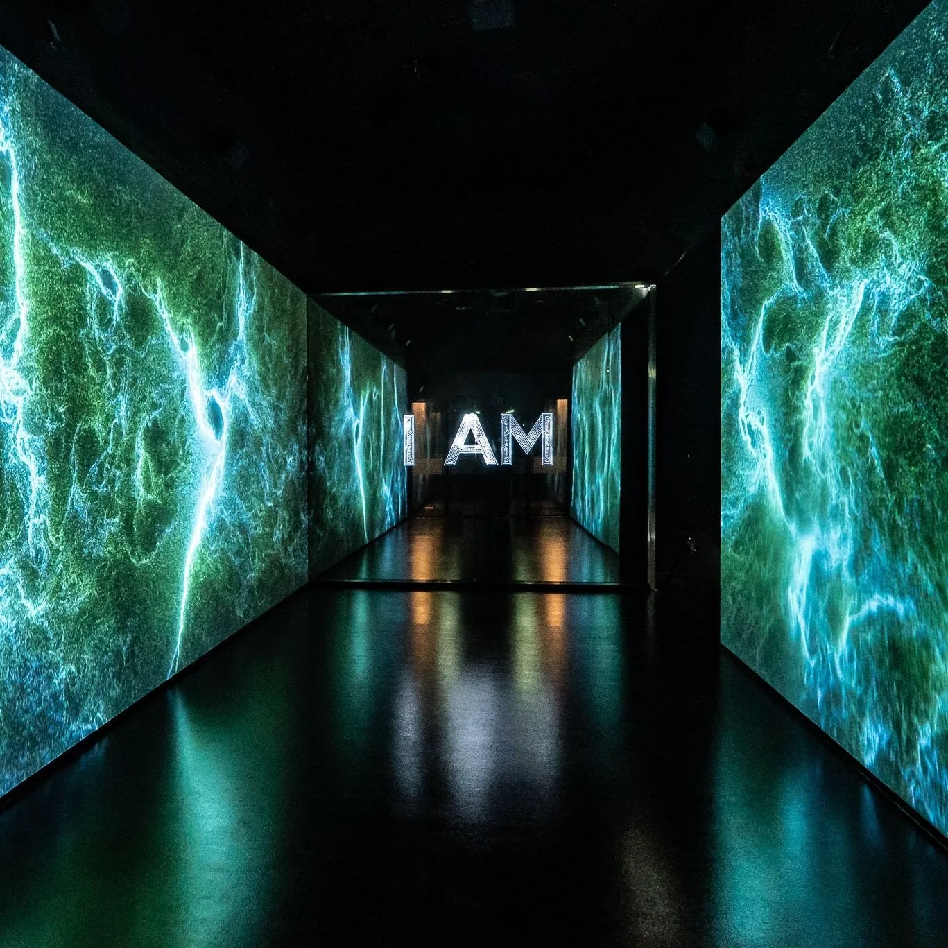 A dark hallway with digital screens displaying flowing water patterns and the words 'I AM' illuminated at the end.