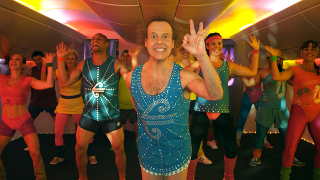 Group of people participating in a lively dance class in a studio with colorful lighting, led by an instructor in the front wearing a blue tank top.