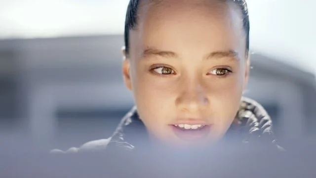 Close-up of a young woman with dark hair tied back, looking at an electronic device with a slight smile.