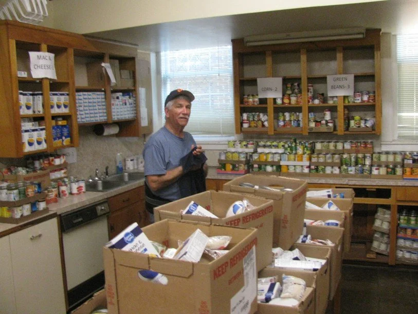 A man smiling while packing food donations in a pantry with cans and boxes of food on shelves.