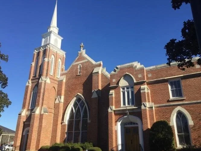 A brick church with a tall steeple against a blue sky, surrounded by trees.