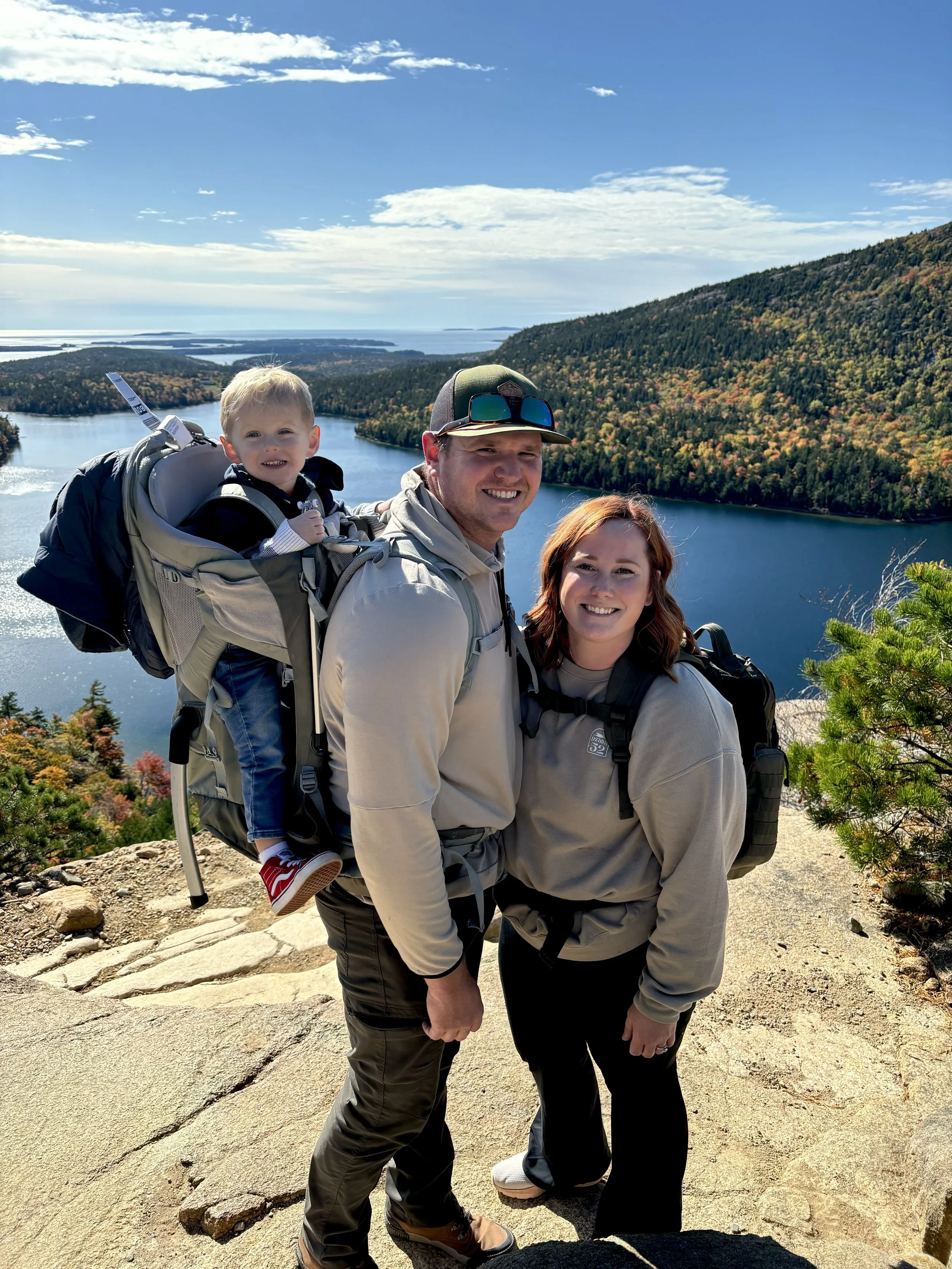 Family of three hiking on a mountain trail with a lake and forest in the background, featuring a father, mother, and young child in a backpack carrier.