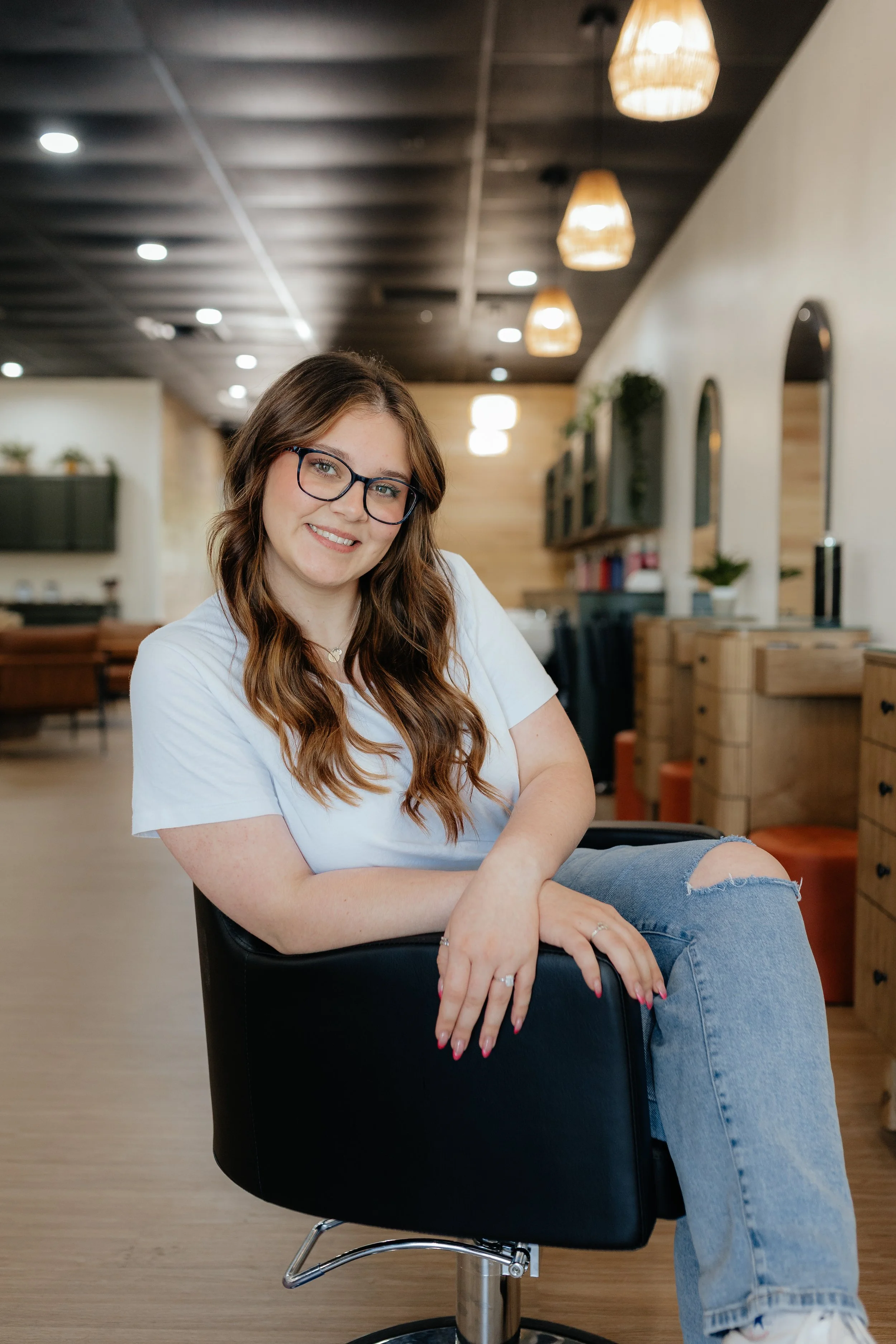 A woman with long wavy brown hair, glasses, and a white t-shirt sitting on a black swivel chair in a modern, well-lit room with wooden cabinets and hanging pendant lights.
