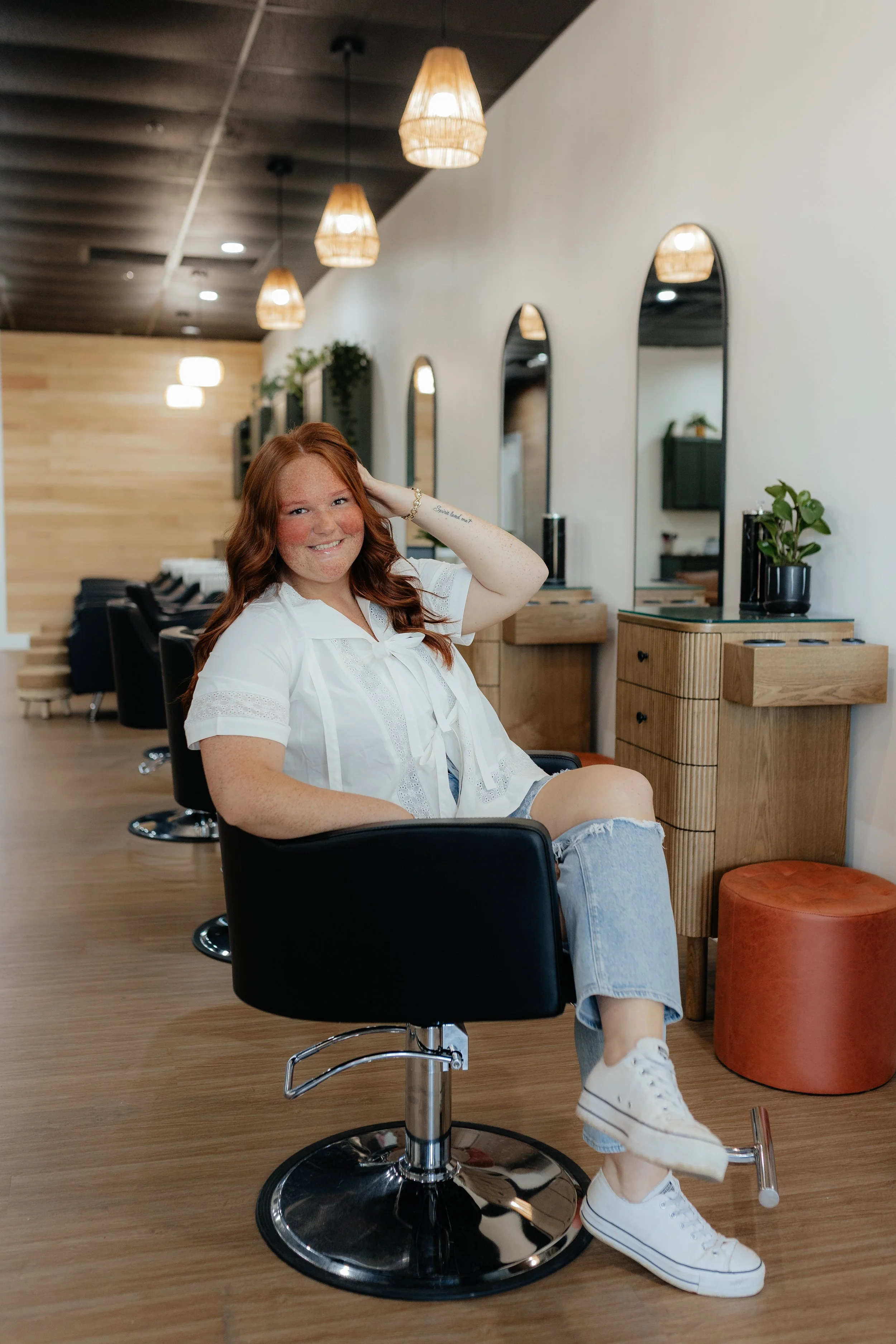 A young woman with red hair, freckles, and a white blouse sitting in a salon chair inside a modern hair salon, smiling and relaxing.