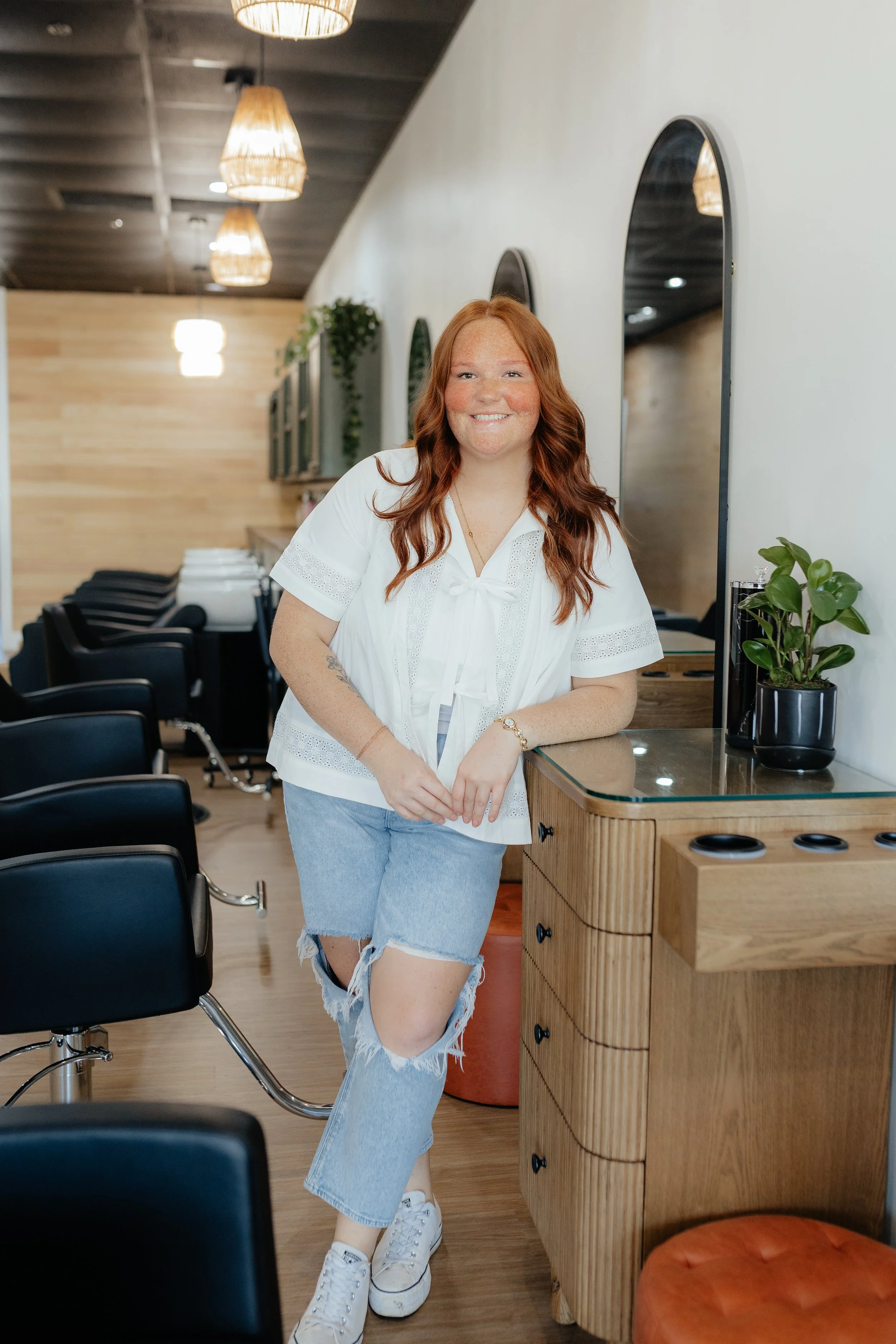 A smiling woman with red hair standing in a modern hair salon, leaning on a wooden counter with a plant on it, and chairs lined up in the background.