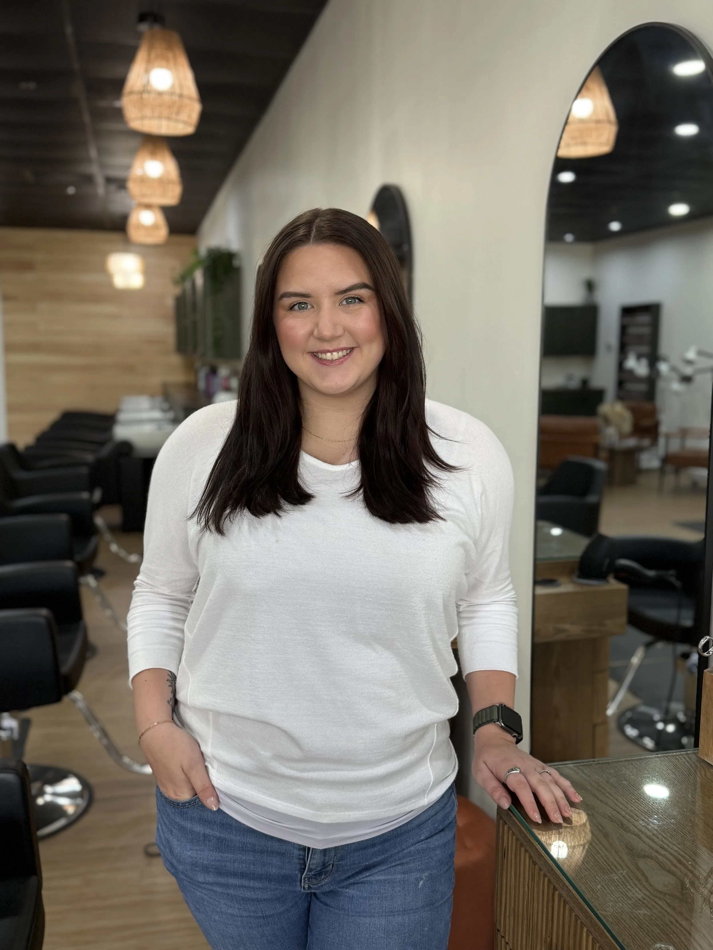 A woman with long dark hair smiling, standing in a salon or beauty salon, wearing a white long-sleeve shirt and jeans, with a watch on her left wrist, standing near a wooden counter with a mirror behind her.