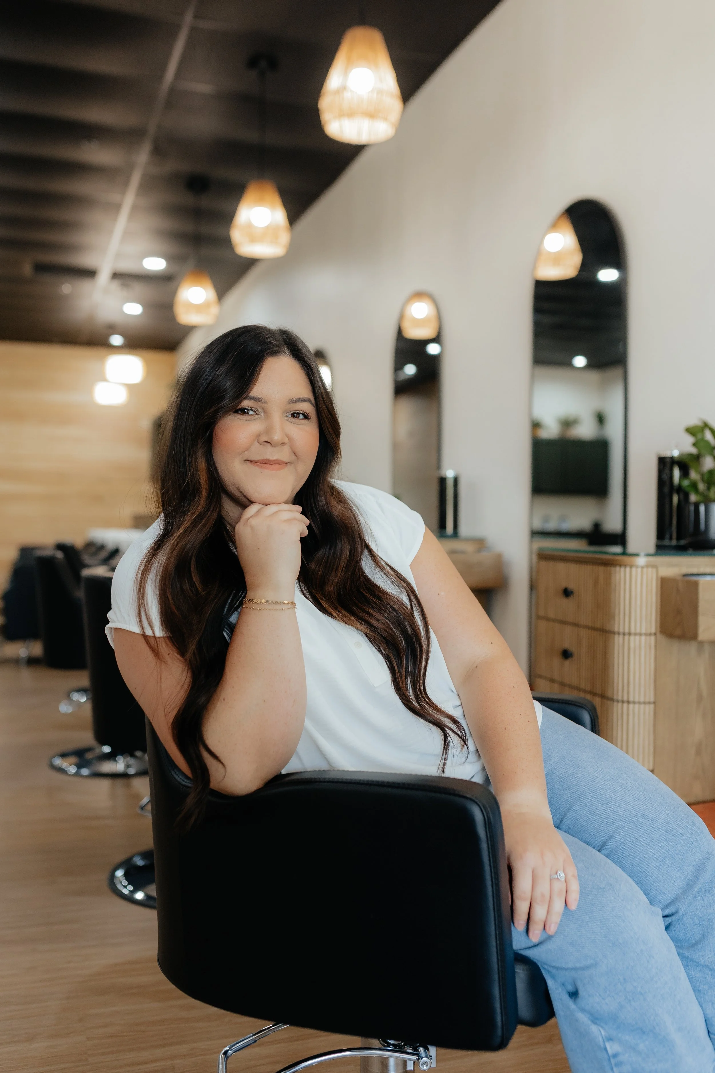 A woman with long brown hair, wearing a white shirt and light blue jeans, sitting on a black salon chair in a modern hair salon, smiling confidently with her chin resting on her hand.