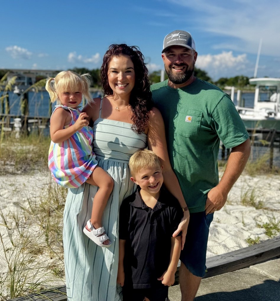 A family of four standing outdoors by a marina with boats in the background, on a sunny day.