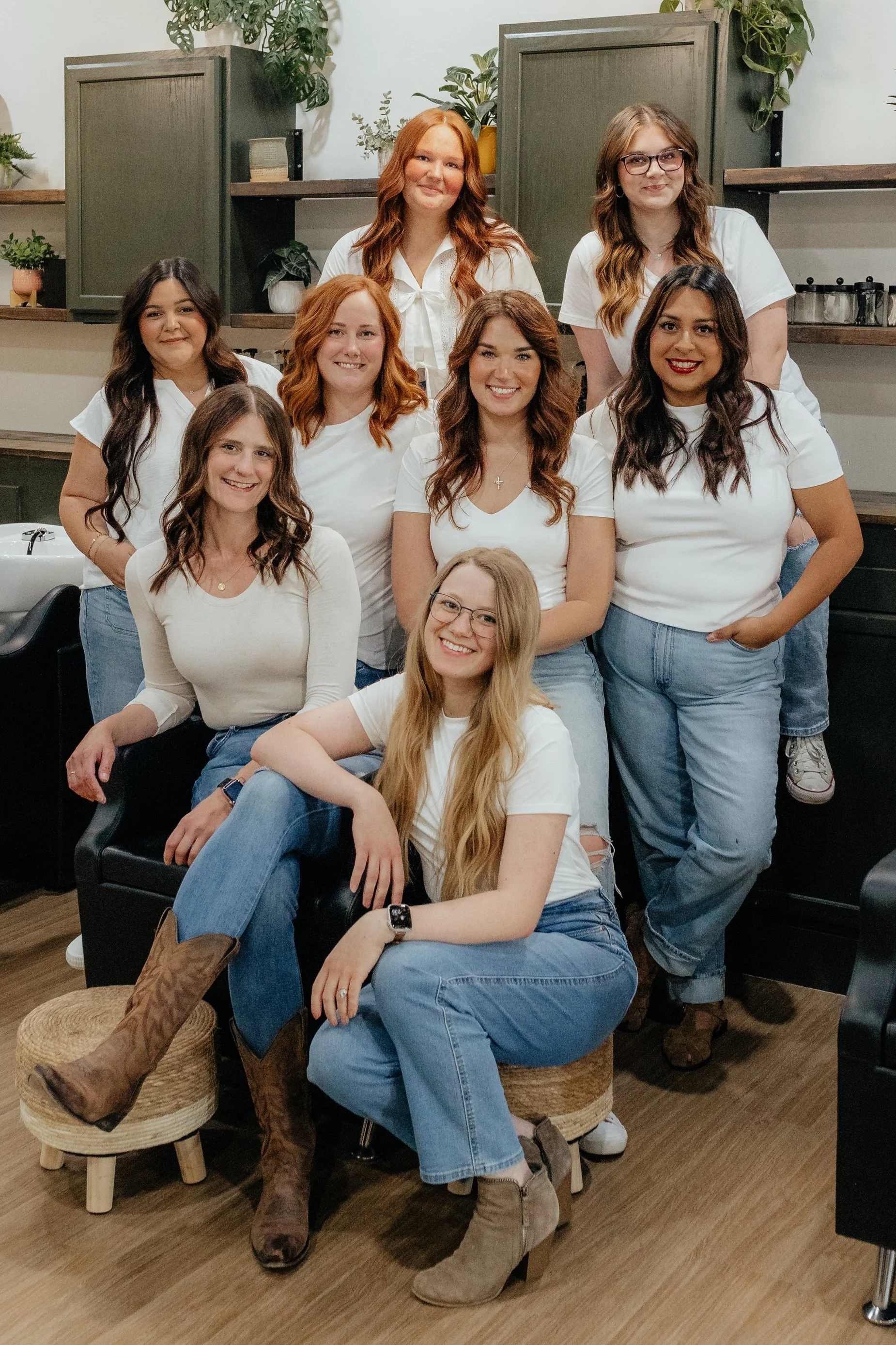 Group of nine women posing together in a room with plants and shelves in the background, all wearing white tops and jeans, smiling at the camera.