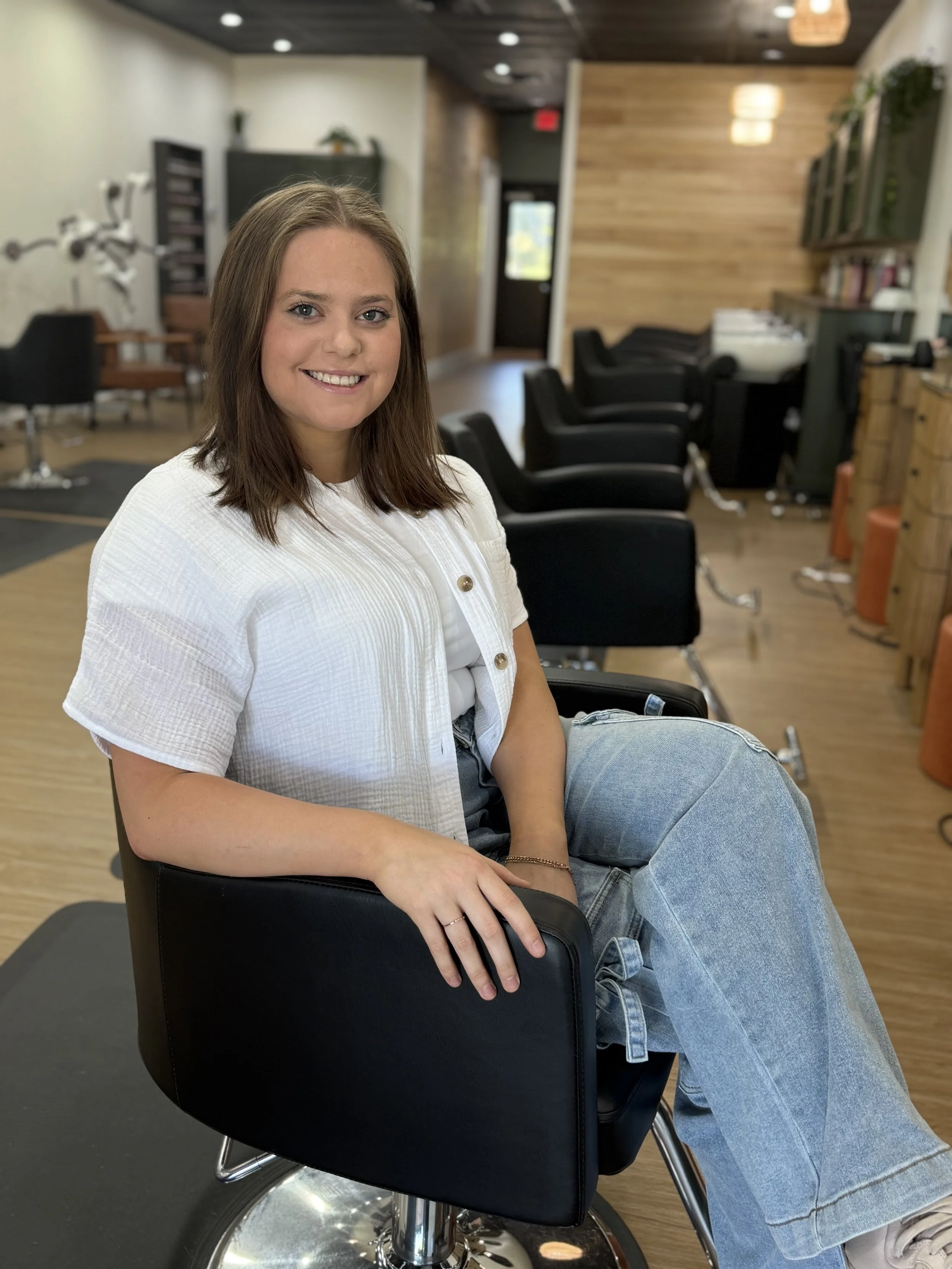 A woman with shoulder-length brown hair sitting in a black salon chair inside a hair salon, smiling at the camera with a background of salon chairs and hair washing stations.