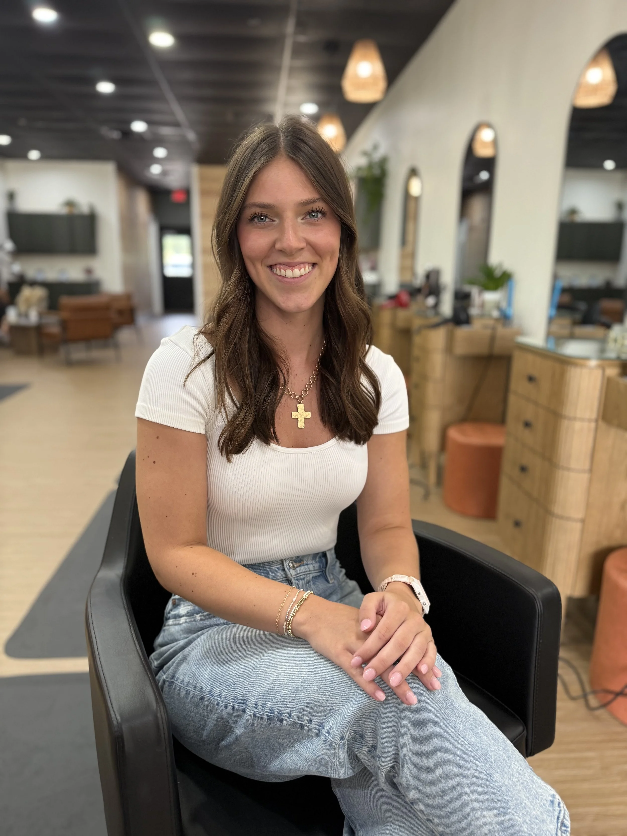 A woman with long brown hair, blue eyes, and a big smile, sitting in a black chair in a modern, warmly decorated cafe or lounge area.