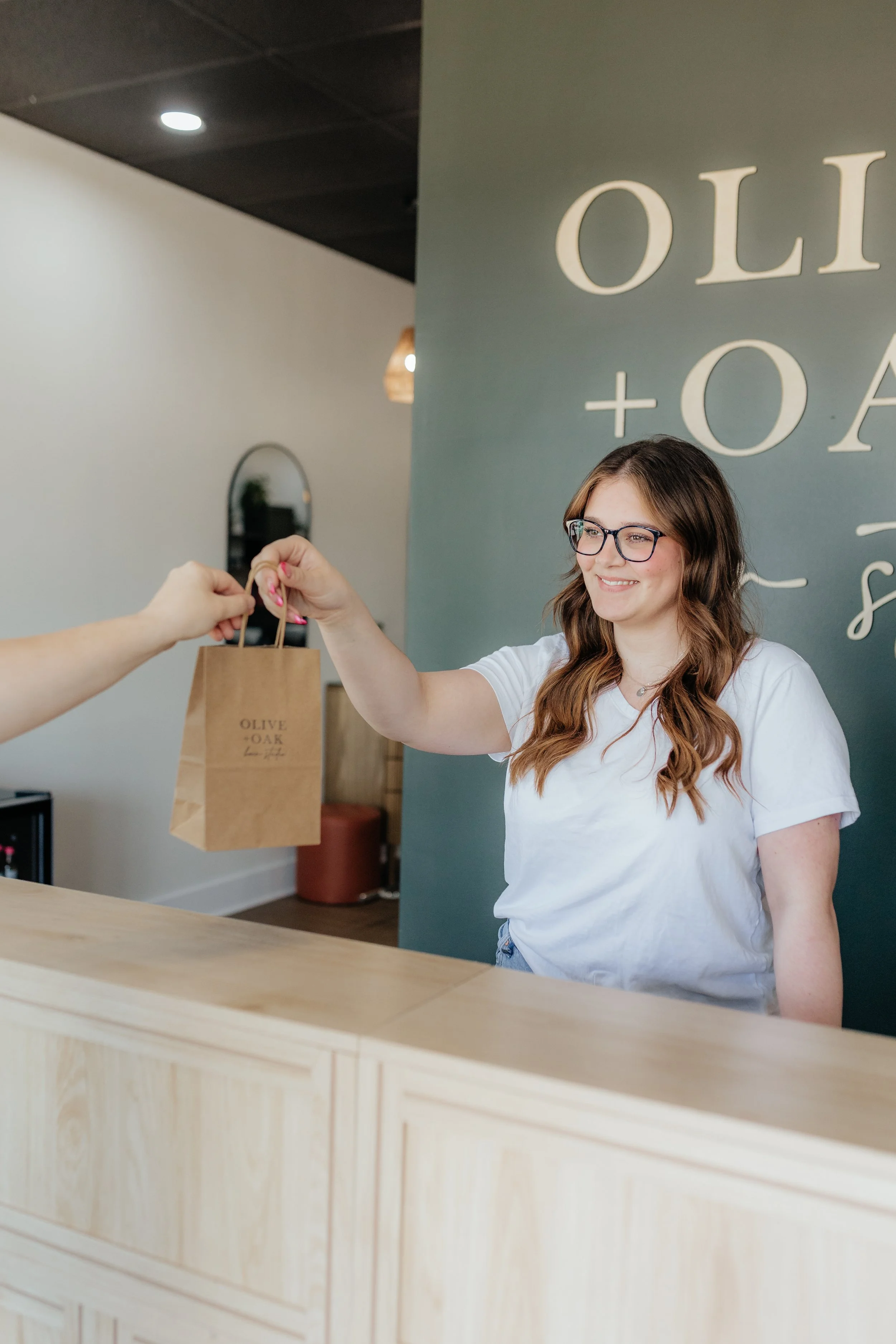A woman with long brown hair and glasses is smiling as she receives a small paper shopping bag from a customer behind a wooden counter in a modern shop.