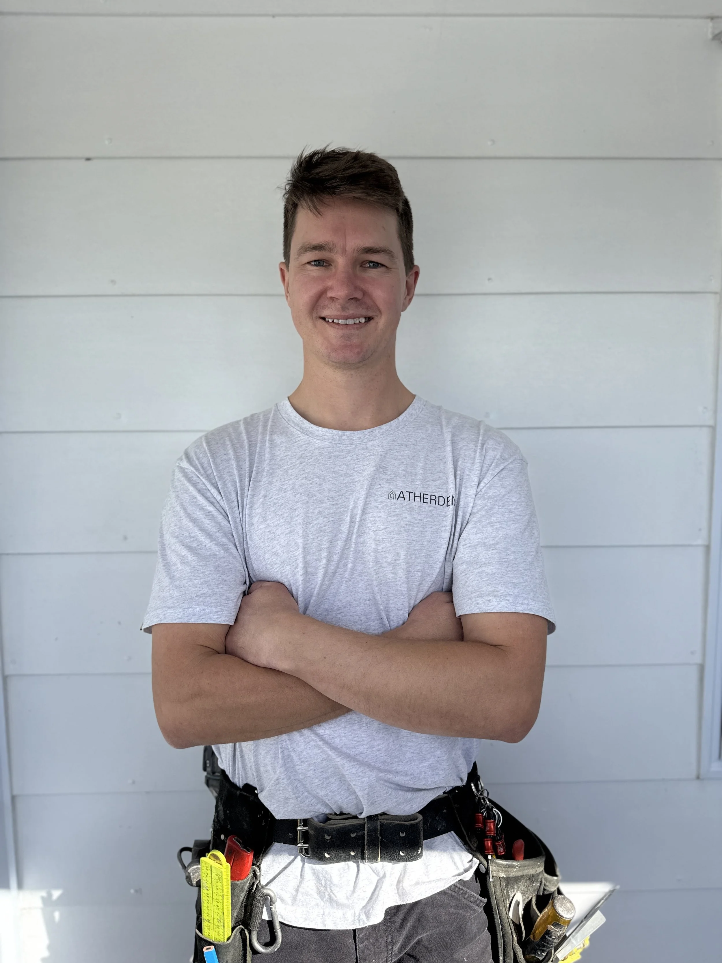 A young man with short brown hair and a smile, standing with arms crossed in front of a white wall, wearing a gray t-shirt and a tool belt with various tools.